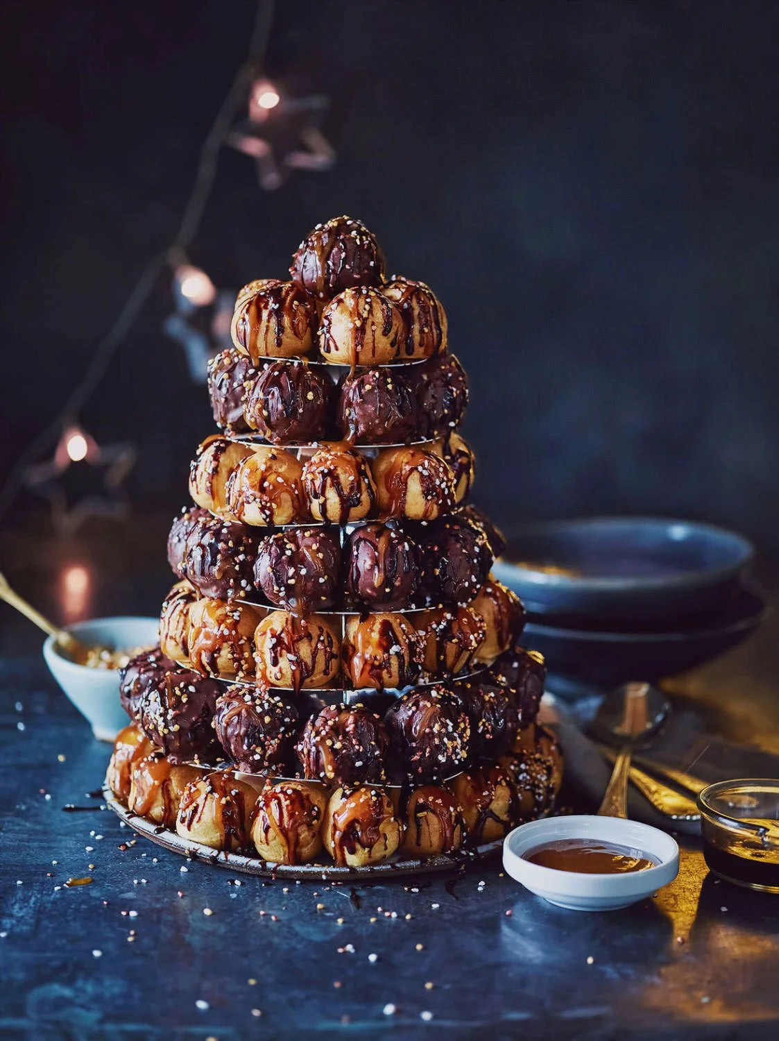A pyramid-shaped display of assorted donuts with chocolate and caramel glazes, drizzled with chocolate syrup and sprinkled with nuts is shown, surrounded by small bowls of sauce and a dark background with string lights.
