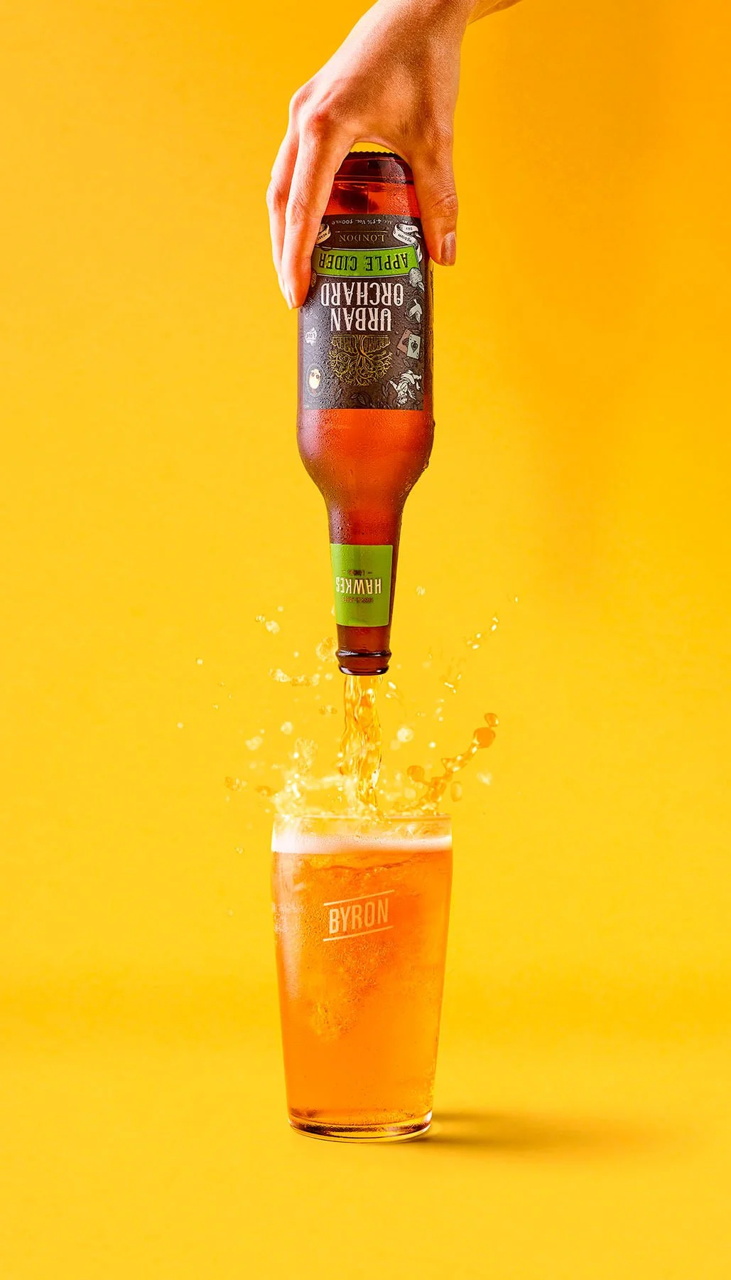 A hand pouring a bottle of beer into a glass against a yellow background.