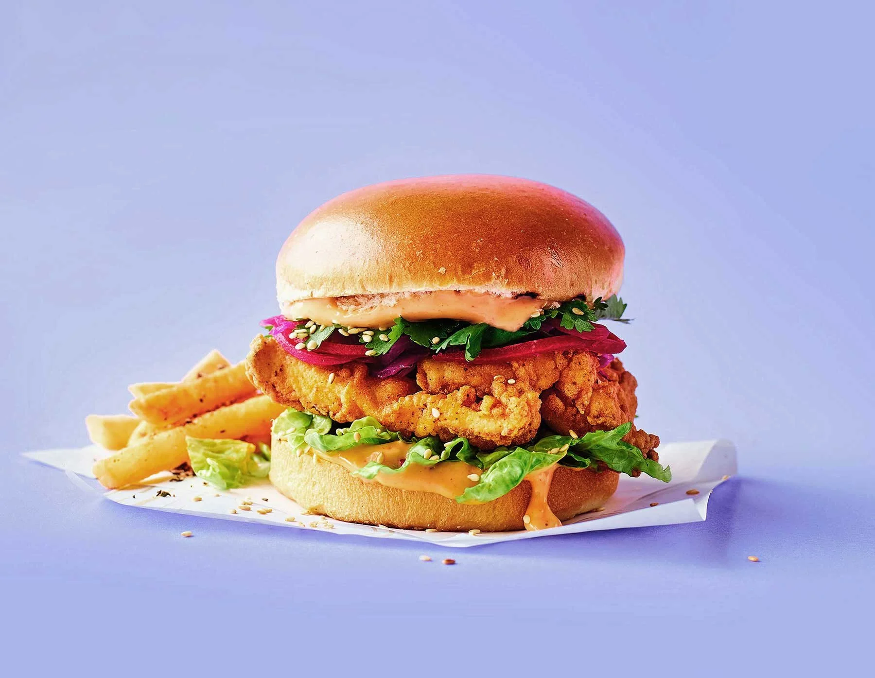Crispy fried chicken sandwich with lettuce, tomato, pickles, spicy mayo, served with French fries on blue background.