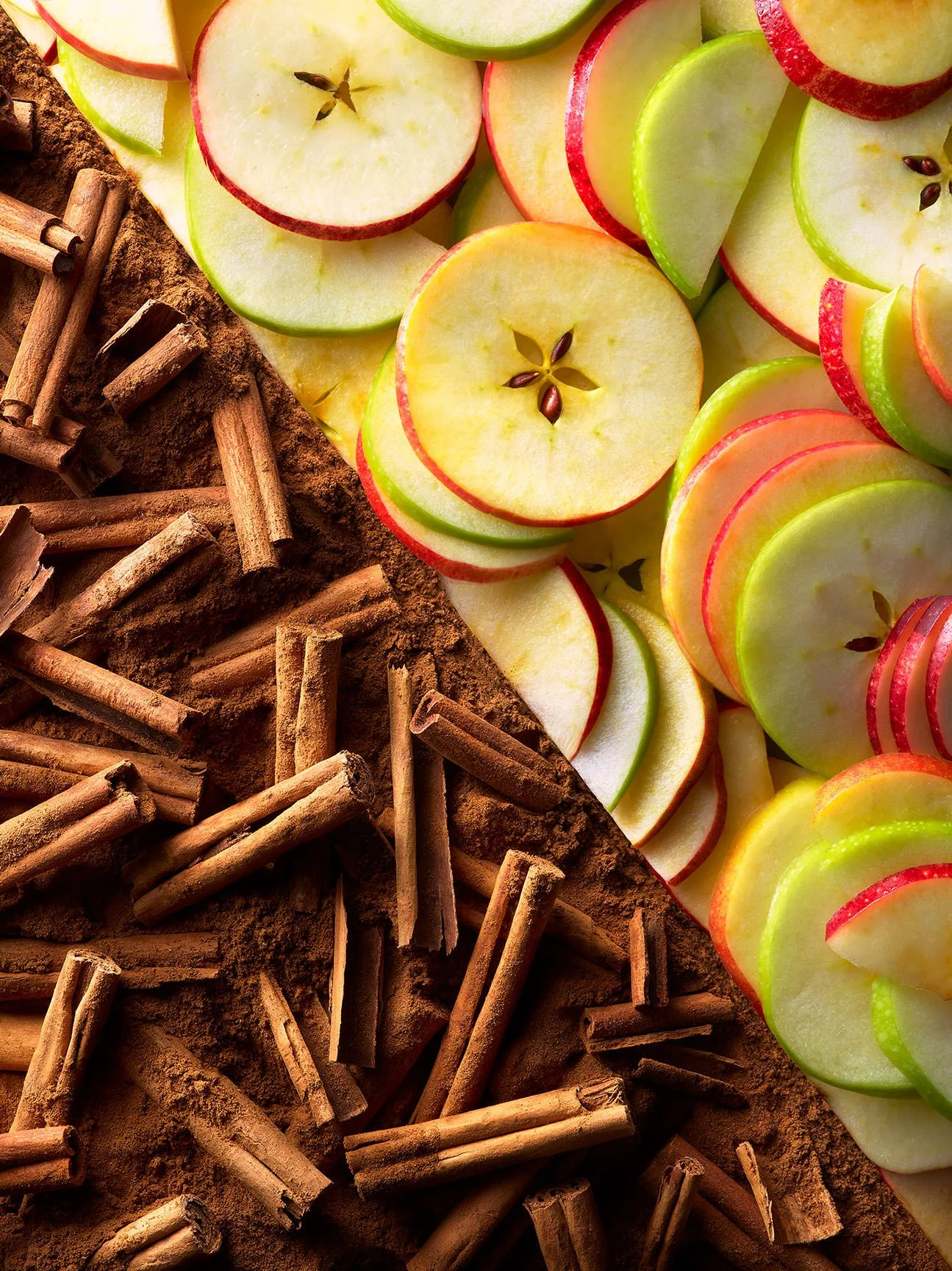 Array of sliced green and red apples on a board with cinnamon sticks and ground cinnamon.