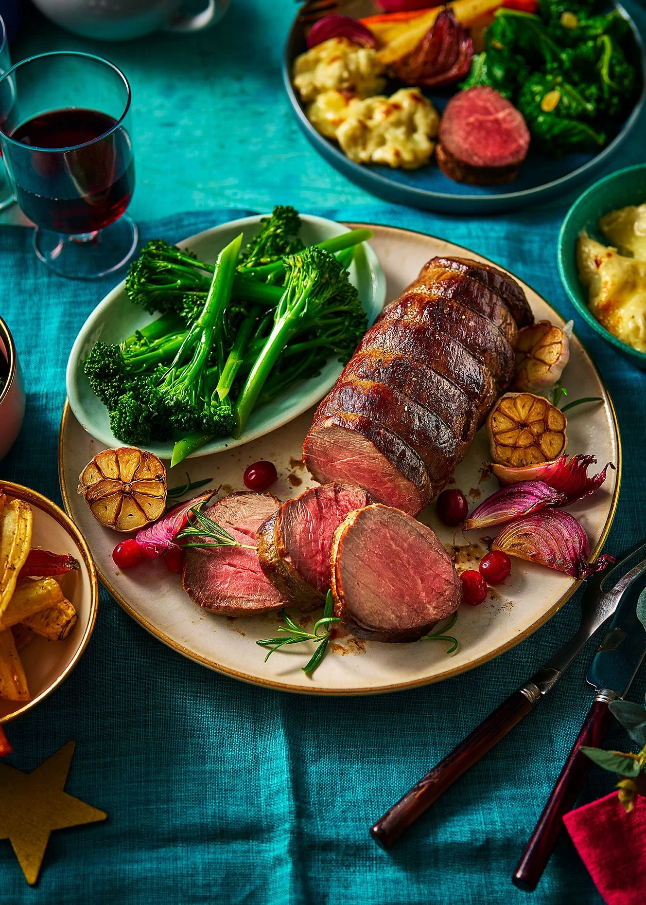 Roast beef sliced on a plate with green broccoli, roasted garlic, and cranberries, surrounded by bowls of mashed potatoes, mashed potatoes with gravy, and another vegetable dish. A glass of red wine is also visible.