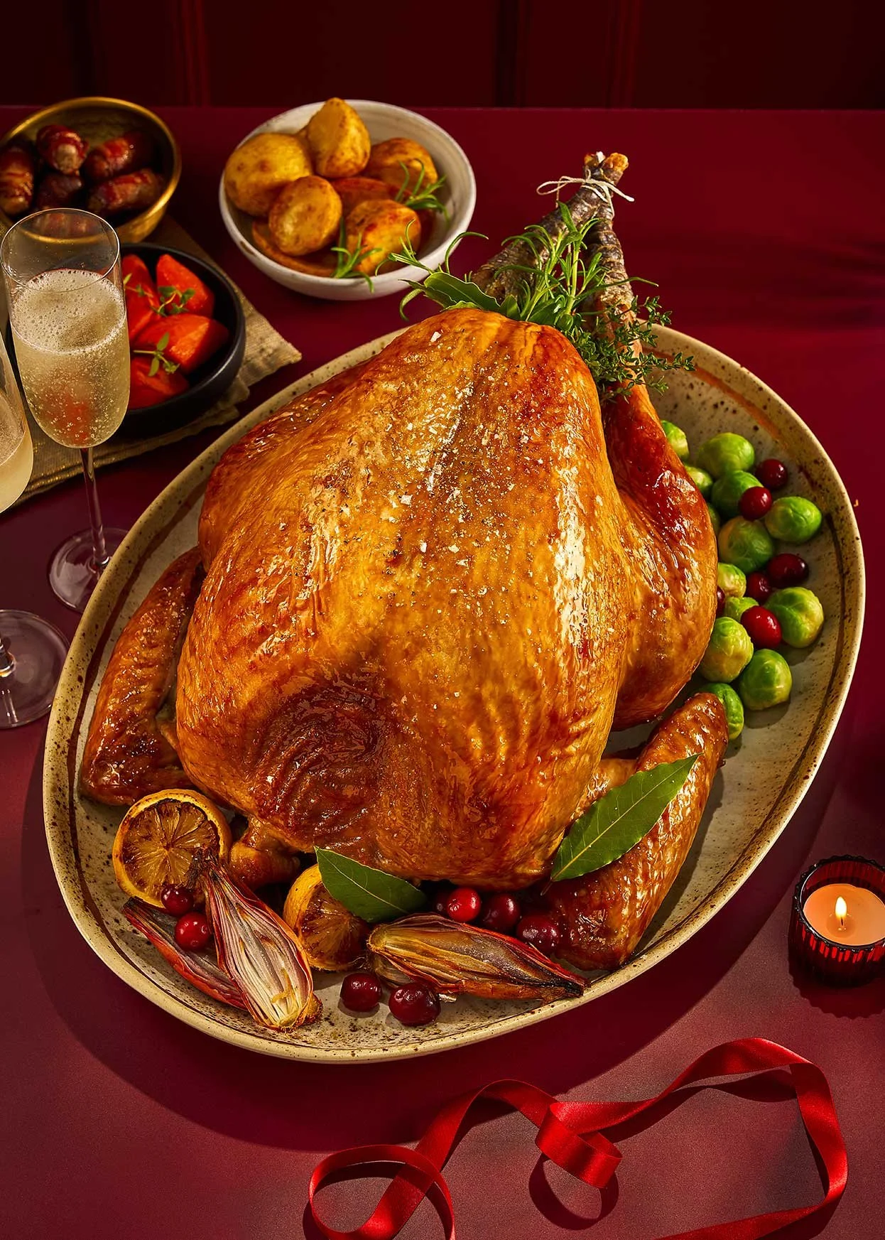A large roasted turkey on a platter garnished with sage leaves, with Brussels sprouts and cranberries around it. Several side dishes and drinks are on the table, including stuffing, mashed sweet potatoes, and glasses of champagne, with a lit candle a