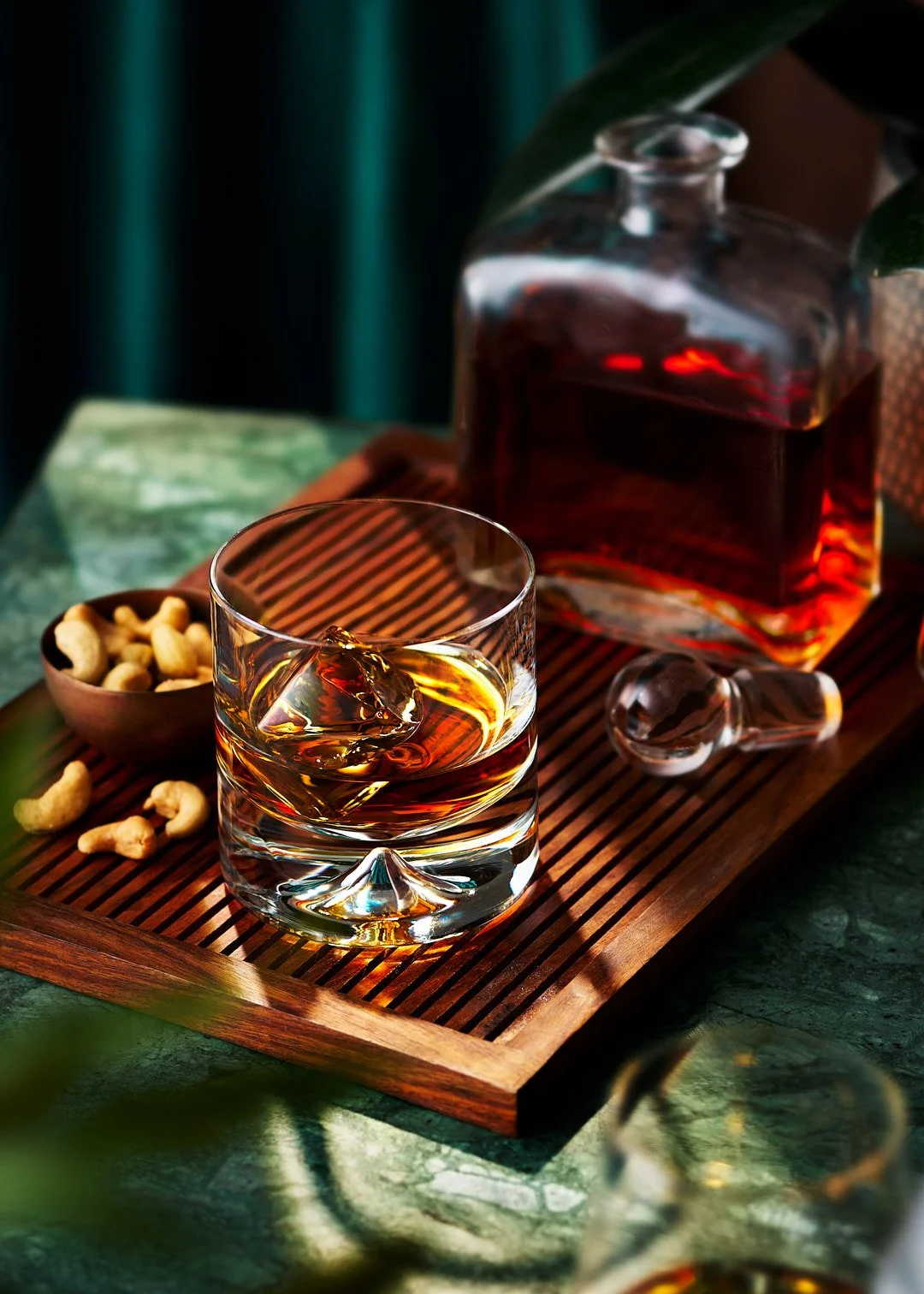 A glass of whiskey with ice on a wooden tray, a decanter filled with whiskey, a small bowl of cashews, and another glass of whiskey in the background.