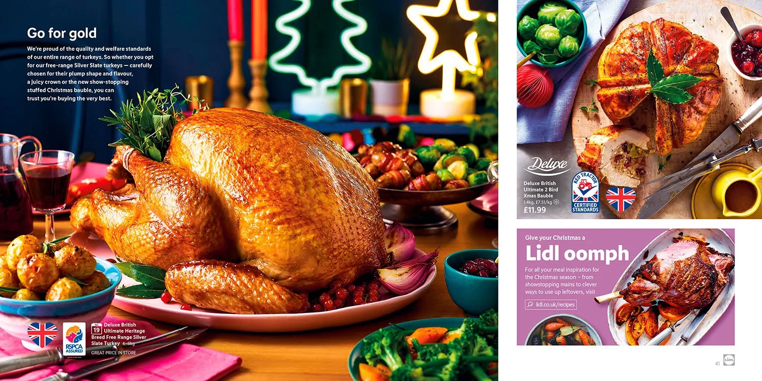 A roasted turkey on a pink platter surrounded by various side dishes including a bowl of stuffing, broccoli, and cranberries. The scene is festive with colorful decorations and illuminated Christmas trees in the background.