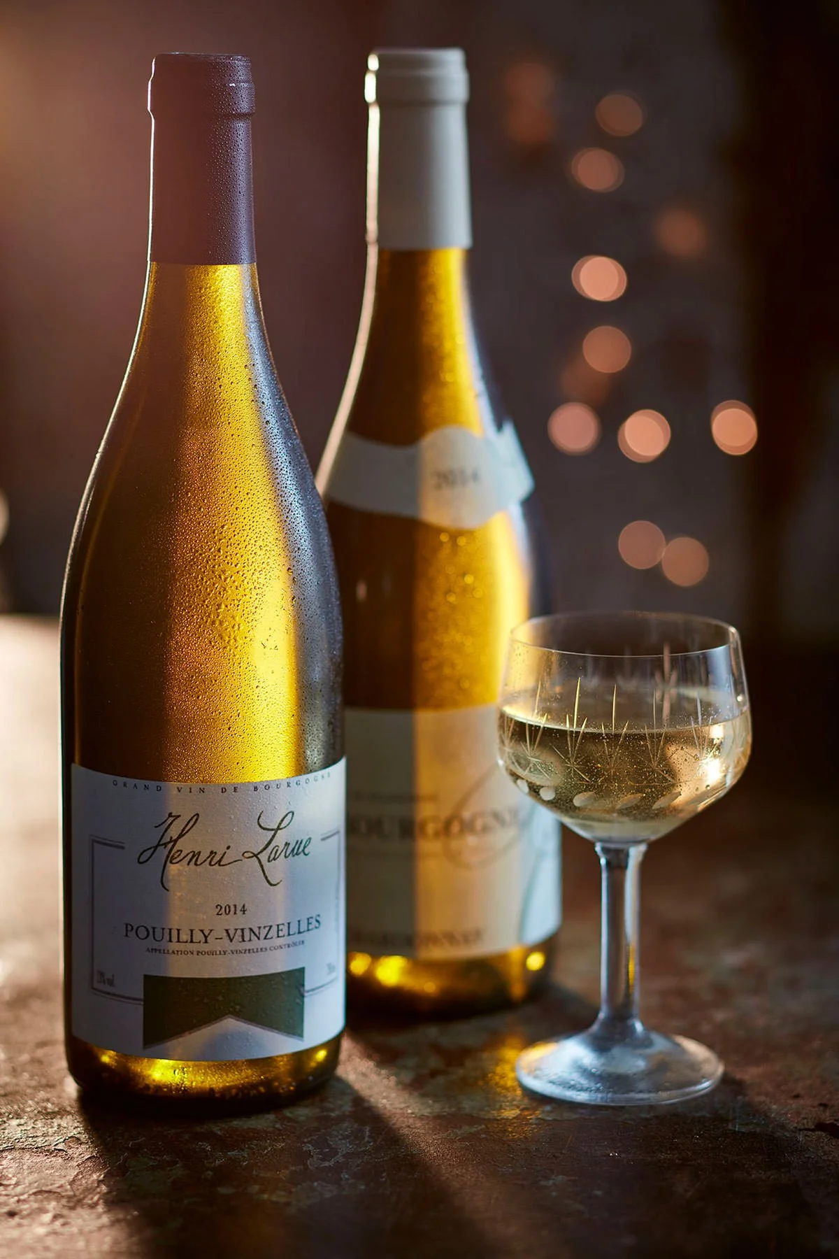 Two bottles of yellow wine or champagne, one in the foreground covered in condensation and a glass with sparkling wine next to them, on a wooden surface with blurred twinkling lights in the background.