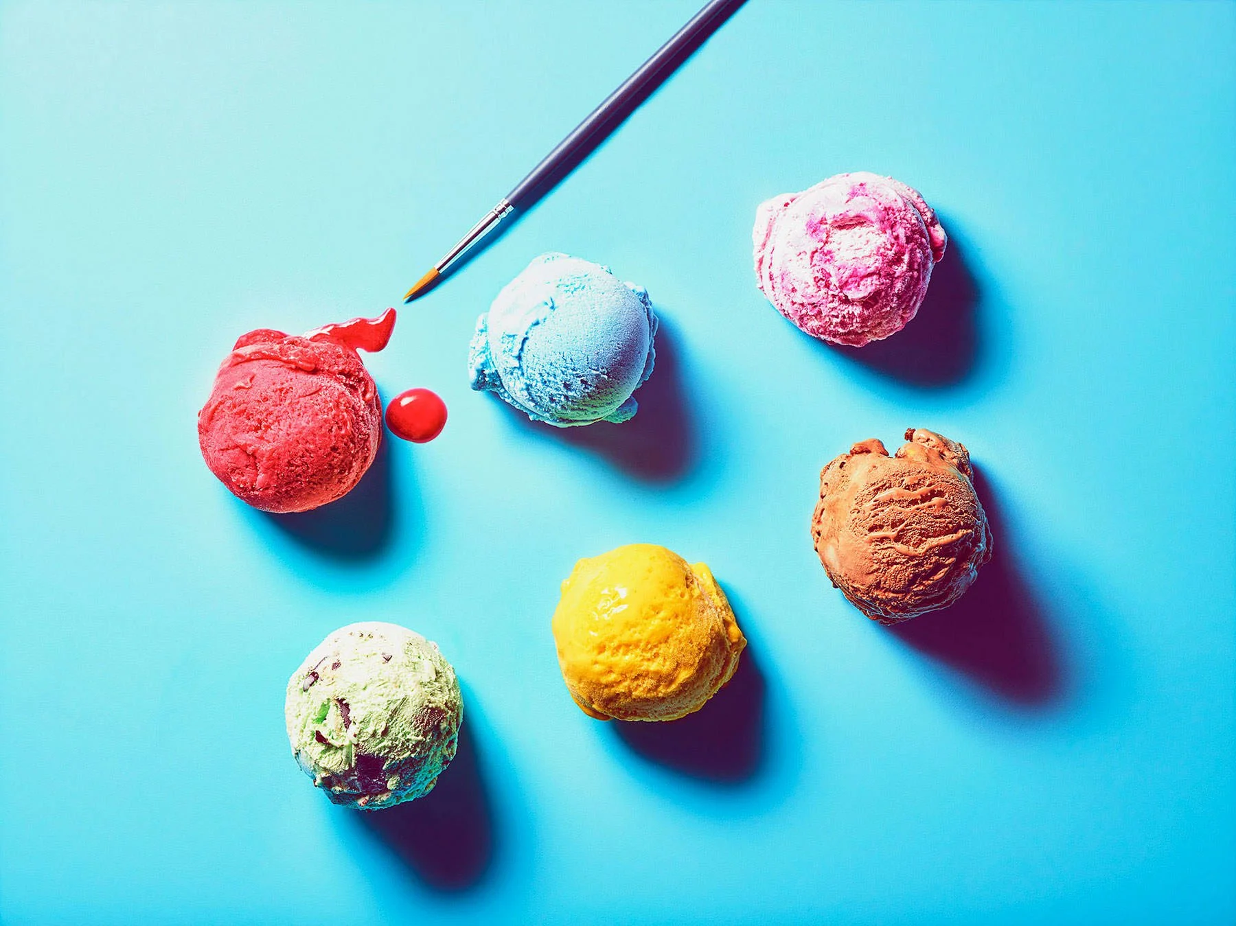 Six colorful scoops of ice cream on a blue surface with a paintbrush and a red cherry.