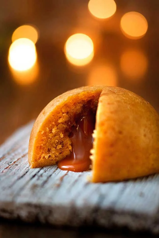A partially broken cookie with caramel filling on a rustic wooden surface, with blurred warm lights in the background.
