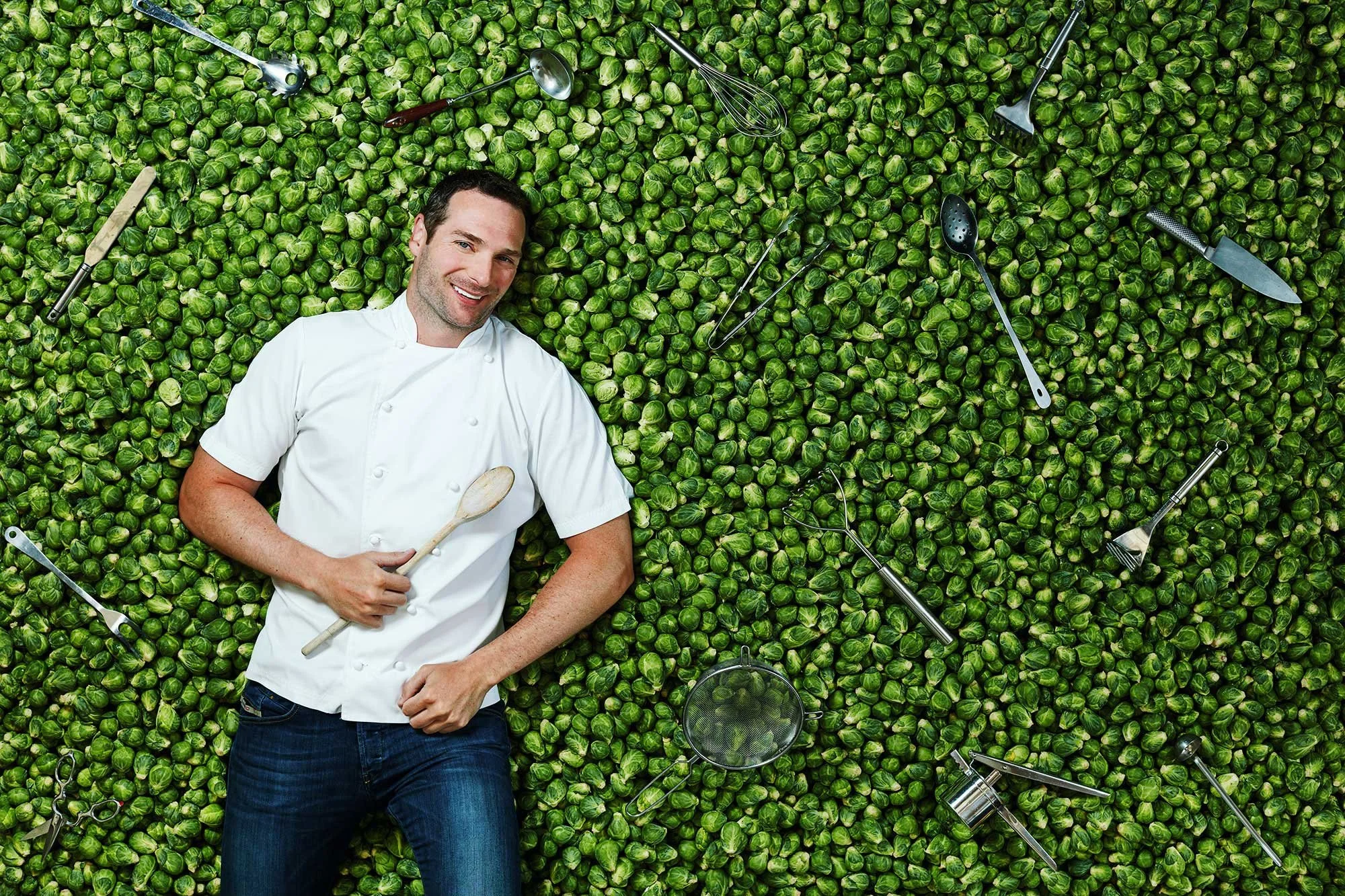 A male chef is lying on a bed of green Brussels sprouts, holding a wooden spoon and smiling at the camera. Surrounding him are various kitchen tools including a whisk, slotted spoon, knife, tongs, sieve, and other utensils.