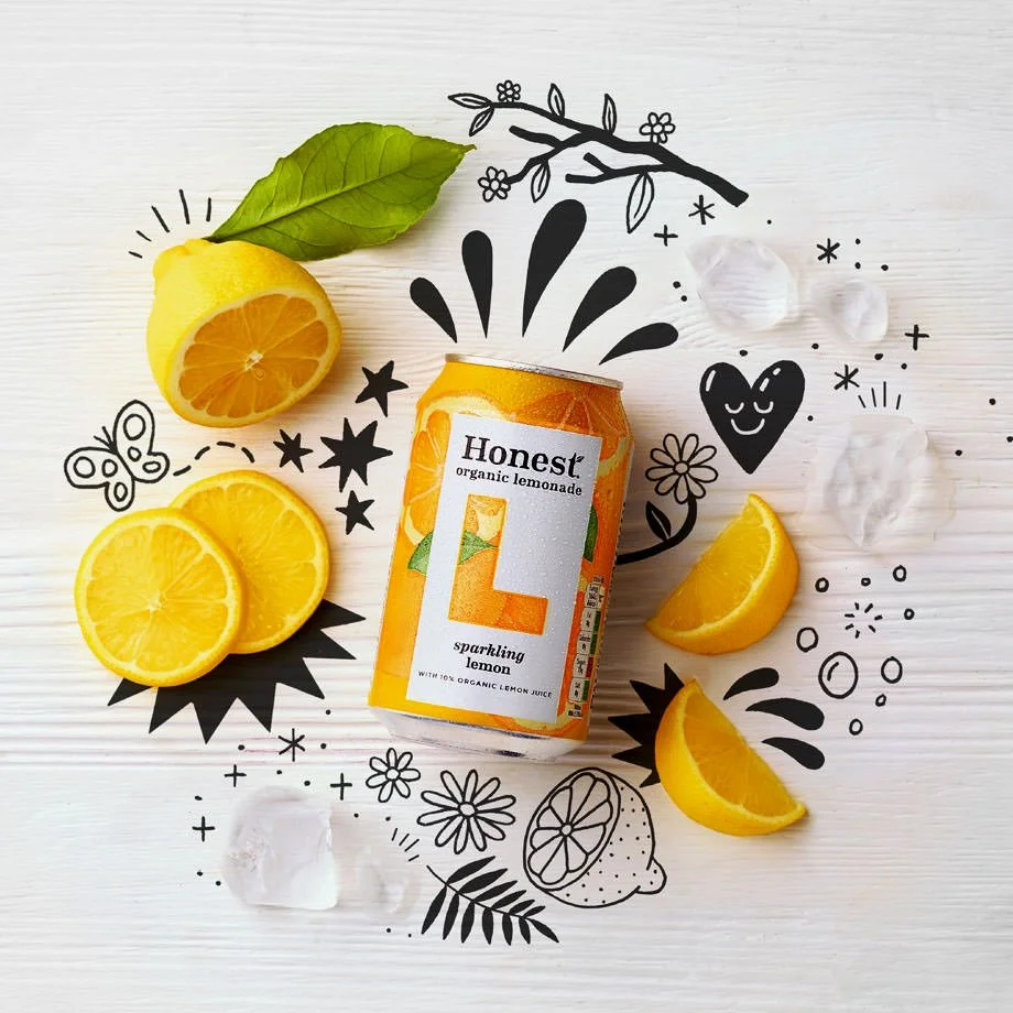 A can of Honest Sparkling Lemonade surrounded by lemon slices, a lemon wedge, ice cubes, a green leaf, and black and white doodles of flowers, a heart, a butterfly, stars, and other decorative elements on a white wooden surface.