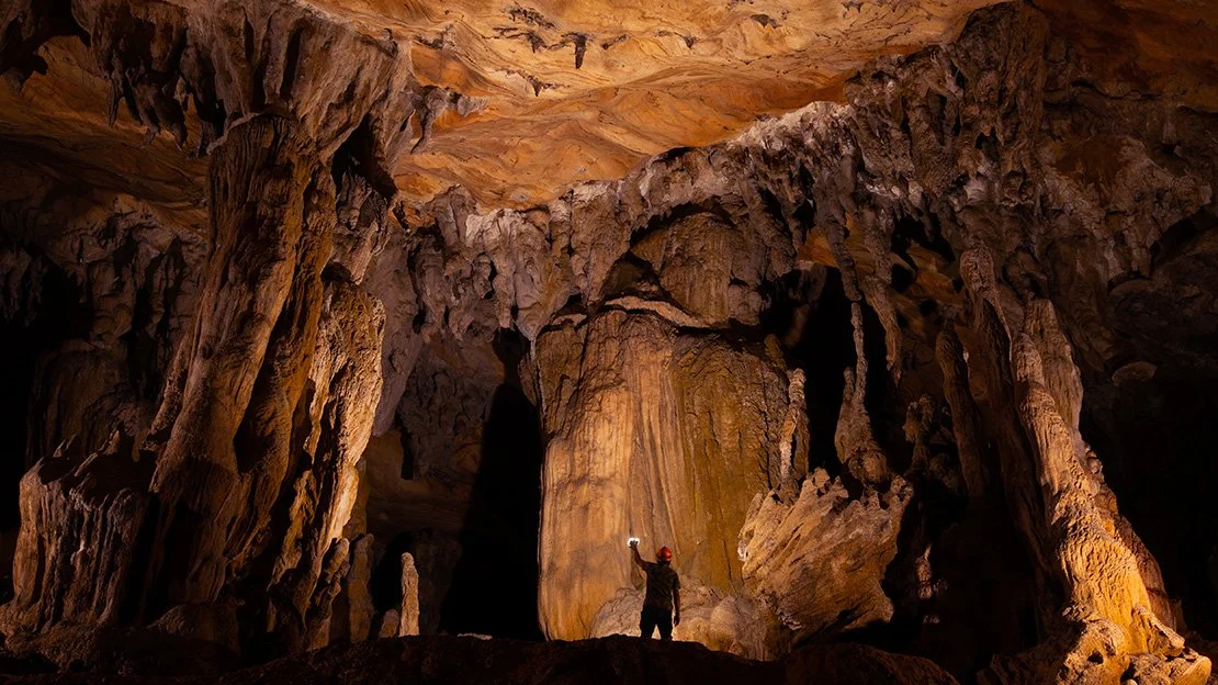 A single figure illuminating a vast cave chamber, revealing rock textures and scale