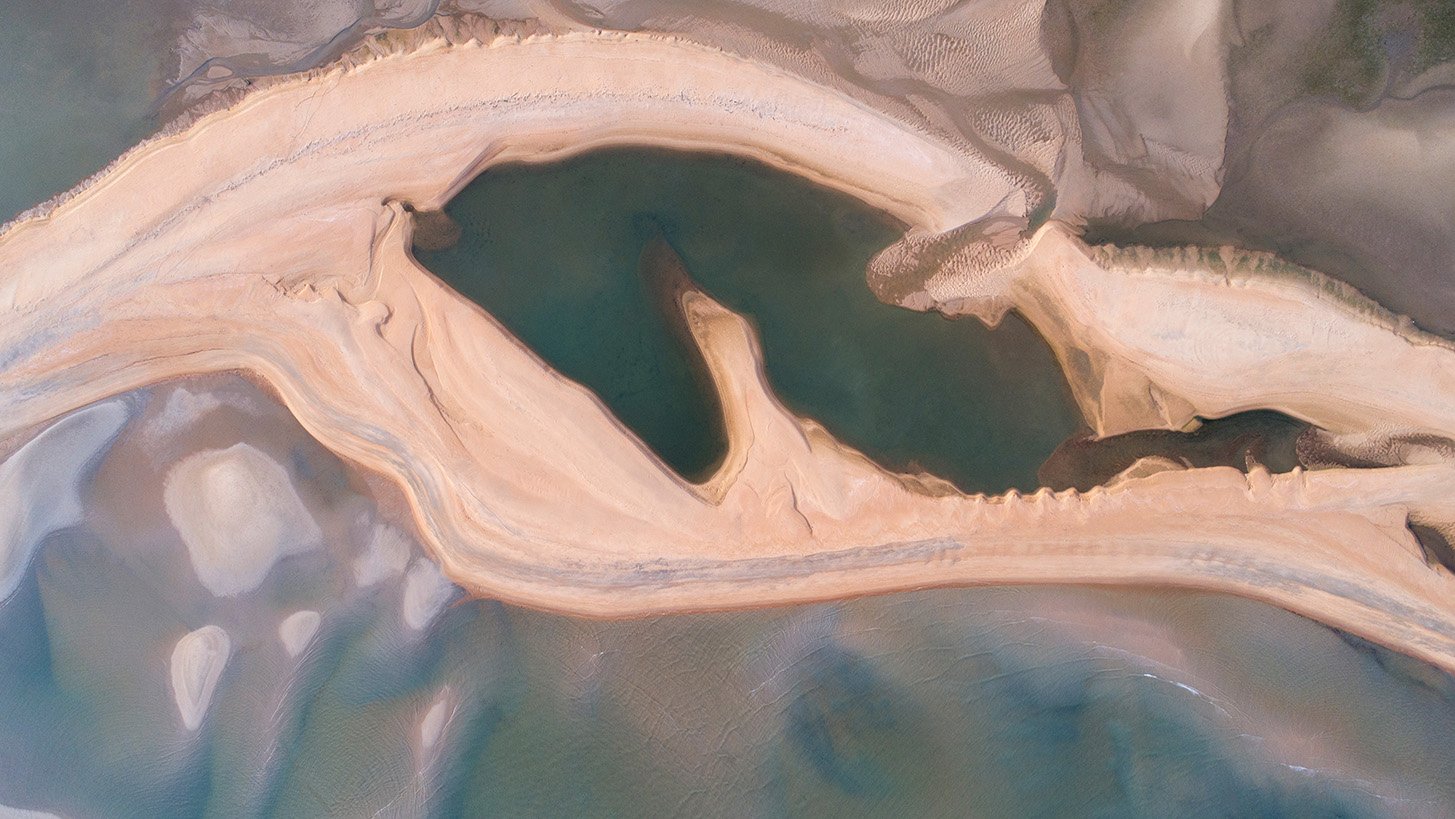 Aerial view of a tidal pool in Chendor, Malaysia, showing pale sand, shallow water, and soft organic shapes formed by the receding tide.