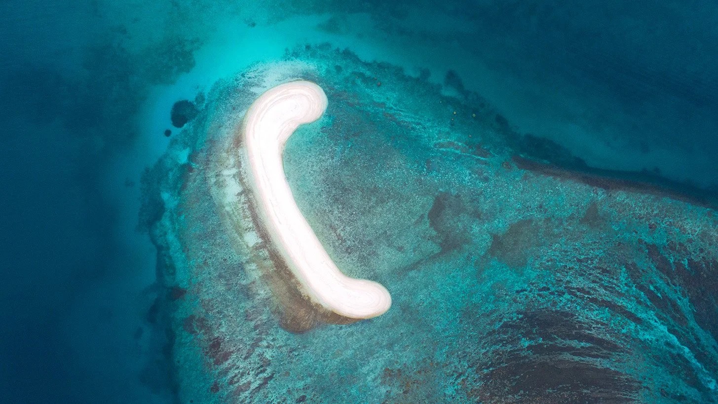 Aerial view of a sandbank near Redang Island with the seabed visible through clear shallow water, revealing subtle tonal shifts and underwater textures.