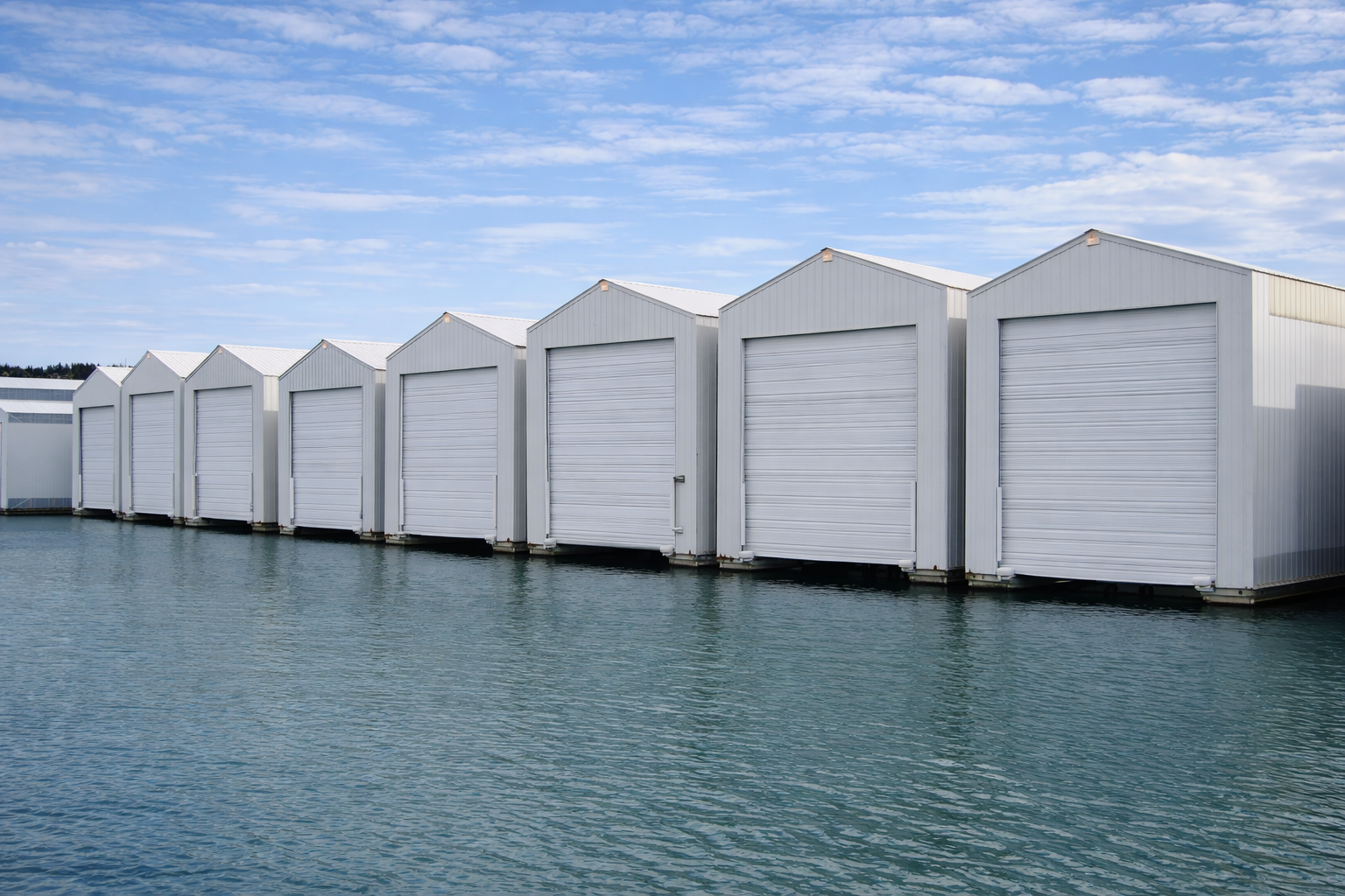 White storage units with closed roll-up doors situated on the edge of a body of water with a partly cloudy sky overhead.
