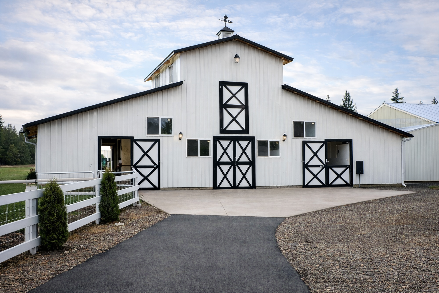 White barn with black trim, multiple small windows, large sliding doors, paved driveway, small trees along white fence.  Raised Center Section Horse Barn in Ferndale, WA