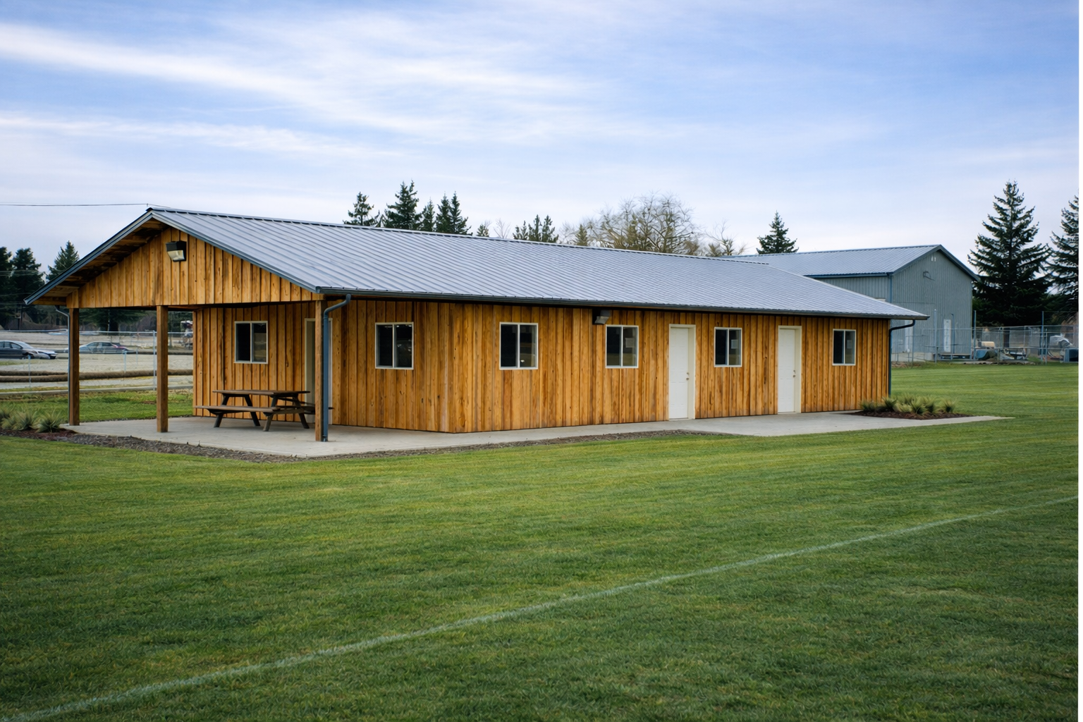 A wooden building with a metal roof on a grassy field, with four windows and two doors, and a small covered porch with a picnic table.