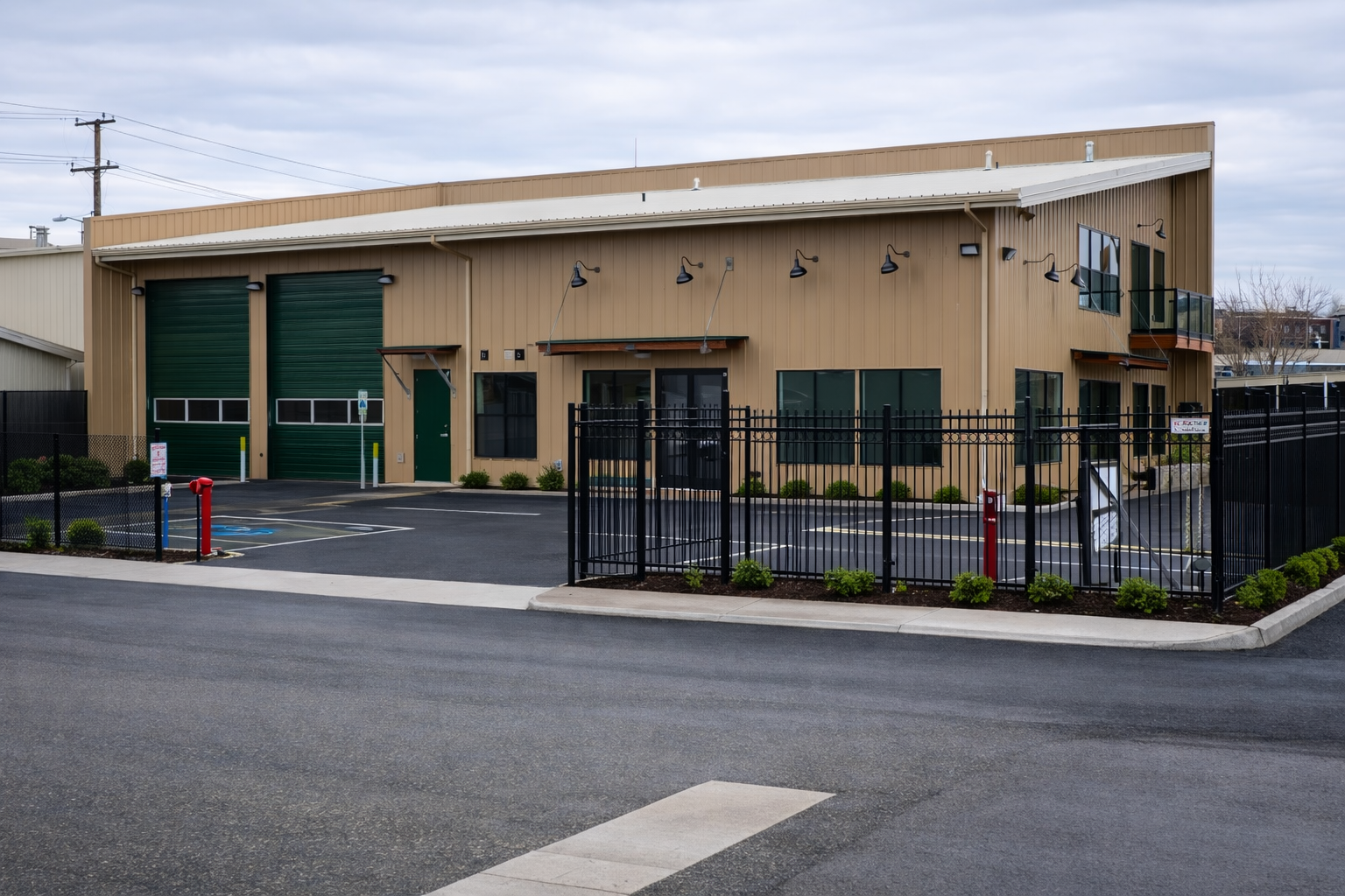 A commercial building with a beige metal exterior, two large green roll-up garage doors, several windows, and a balcony. The building is surrounded by a black metal fence with small shrubs and a parking lot with marked spaces.