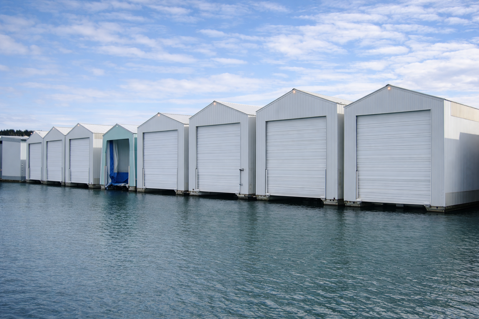 Row of white boat storage sheds on a body of water under a partly cloudy sky.