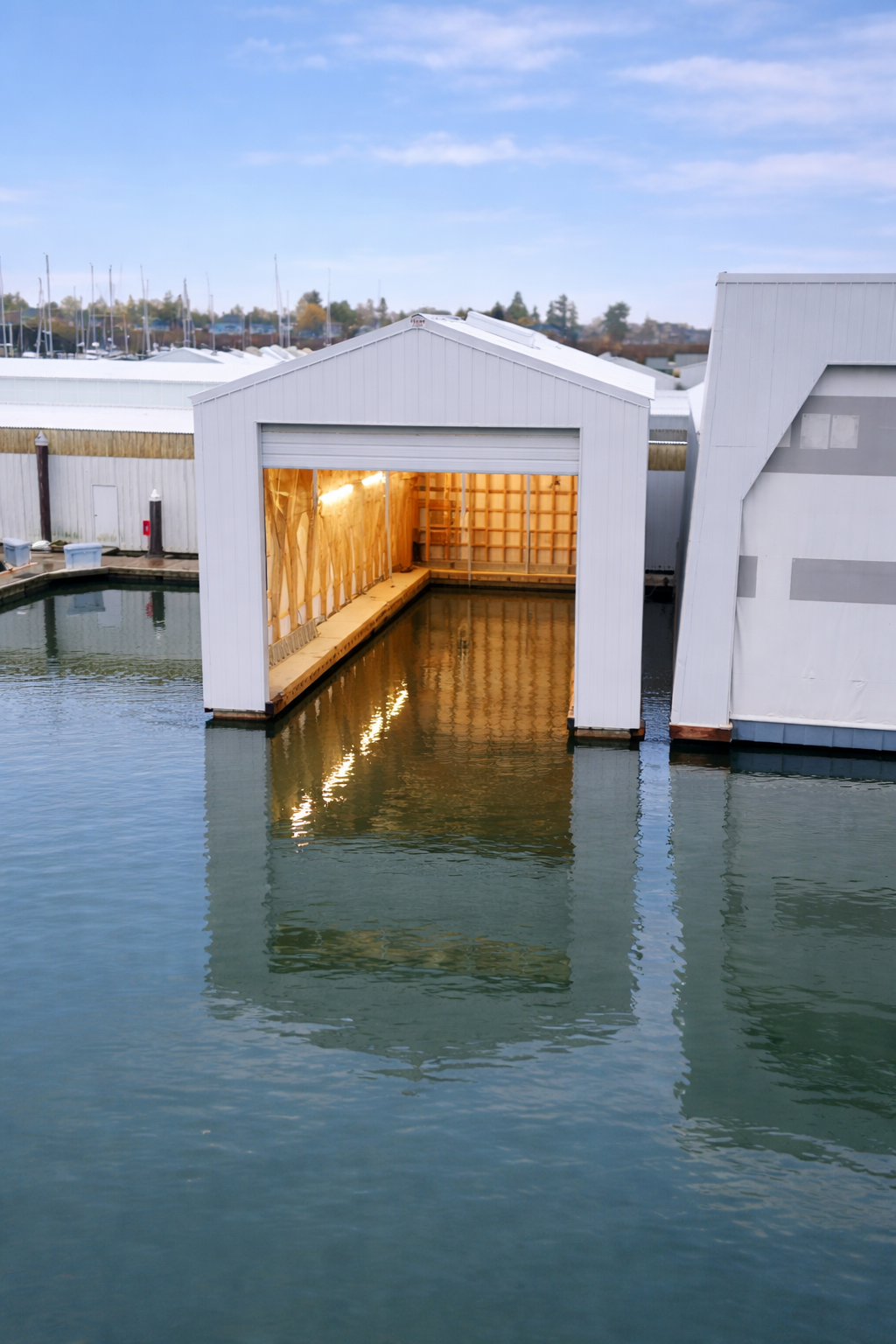 A boat garage with its door open, built on the water, revealing a wooden interior with lighting. Surrounding the garage are calm water and other white industrial buildings, with a background of a cloudy sky and a marina with sailboats.