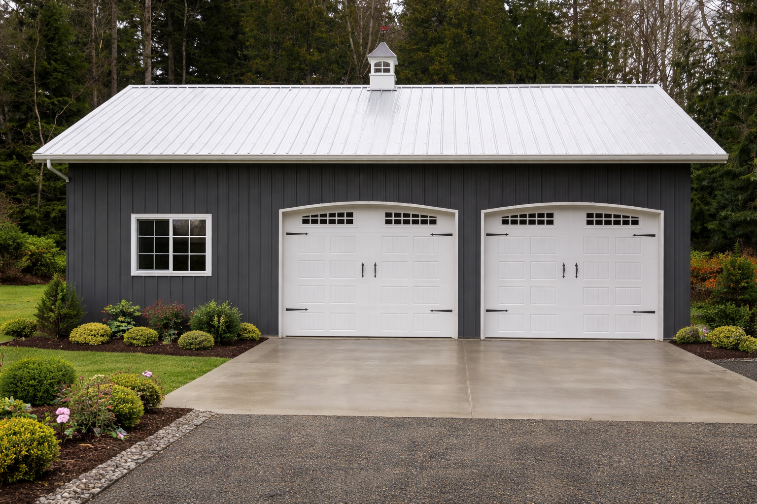 A gray garage with two white doors and black hardware, a single window on the left, surrounded by green bushes and a landscaped yard, with a concrete driveway leading to the garage, and a wooded background. Post Frame Building in Bellingham.