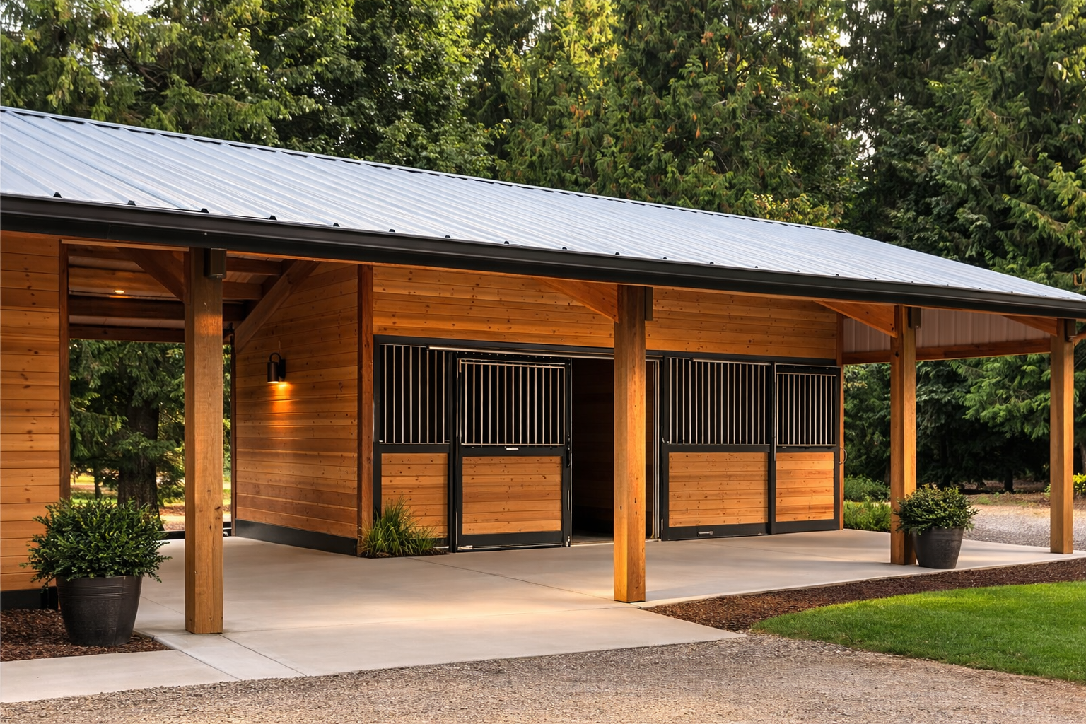 A wooden horse stable with metal roof, black metal gates, outdoor lighting, potted plants, concrete pathway, green lawn, and trees in the background.