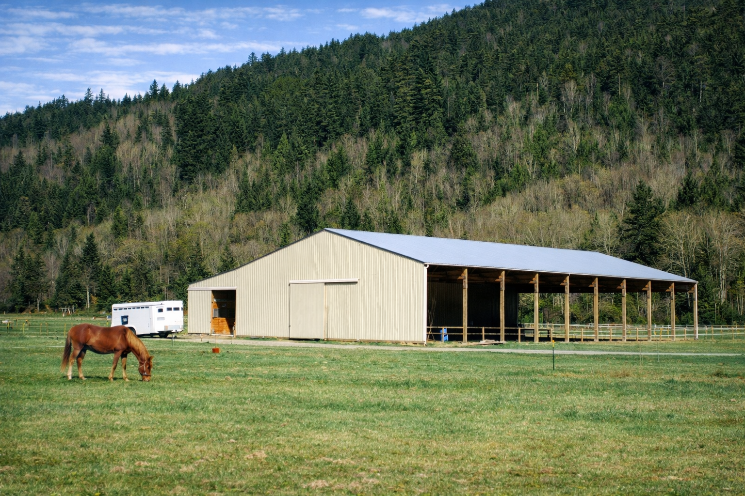 A brown horse grazing on green grass in front of a large beige barn with a metal roof, surrounded by a fenced pasture, with a white horse trailer nearby and a hillside covered in trees in the background under a partly cloudy sky.