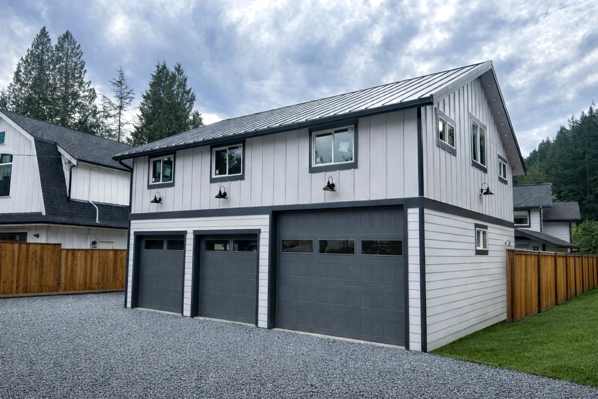 Two-story modern barndaminum house with a gray garage door, white siding, black trim, and large windows, surrounded by a wooden fence and green lawn, with a cloudy sky overhead. barndominium in Whatcom County.
