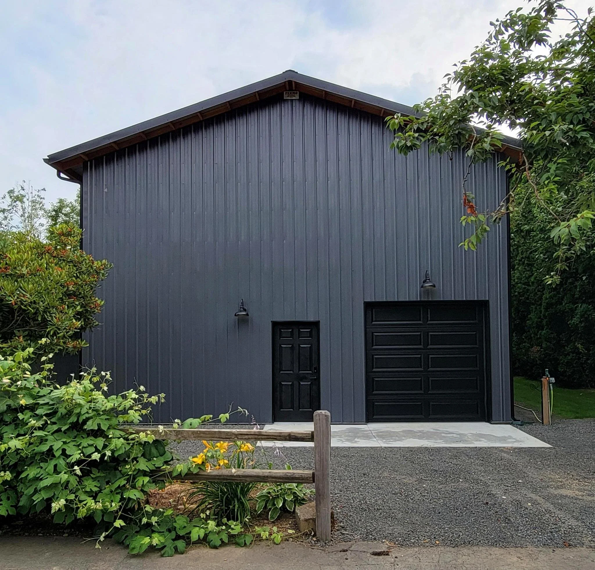 A modern, dark gray metal garage with a black door and a separate black entry door, surrounded by green plants and trees.