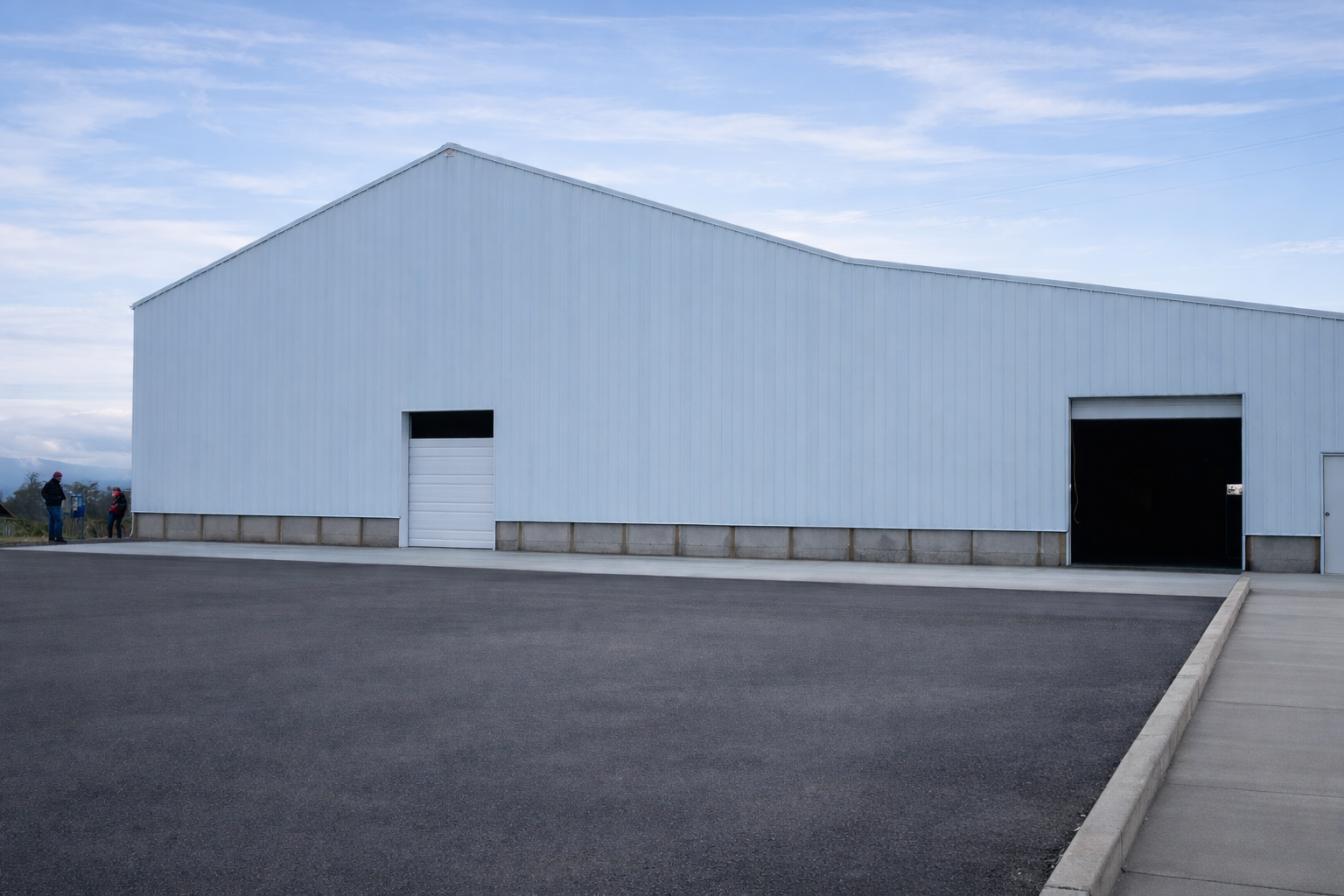 A large blue warehouse with a metal exterior, featuring two large garage doors, with a concrete pathway and a wide paved area in front.