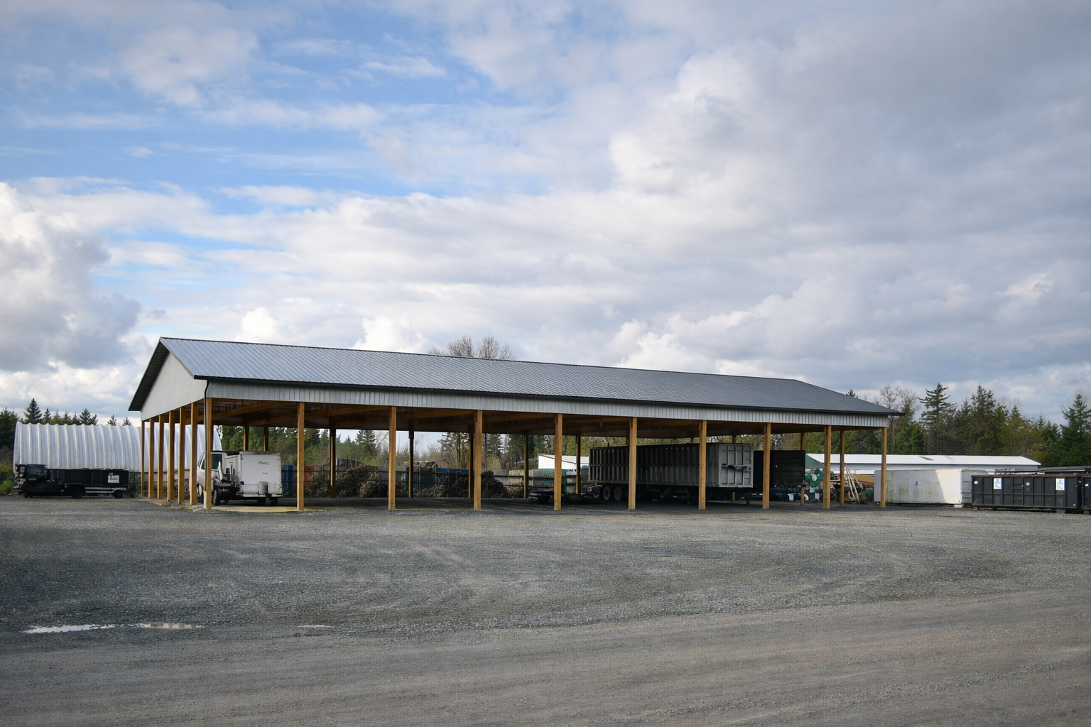 Metal storage shed with a sloped roof and open sides, situated on a gravel lot, with various trailers and equipment underneath and around it, against a partly cloudy sky and distant trees in the background.
