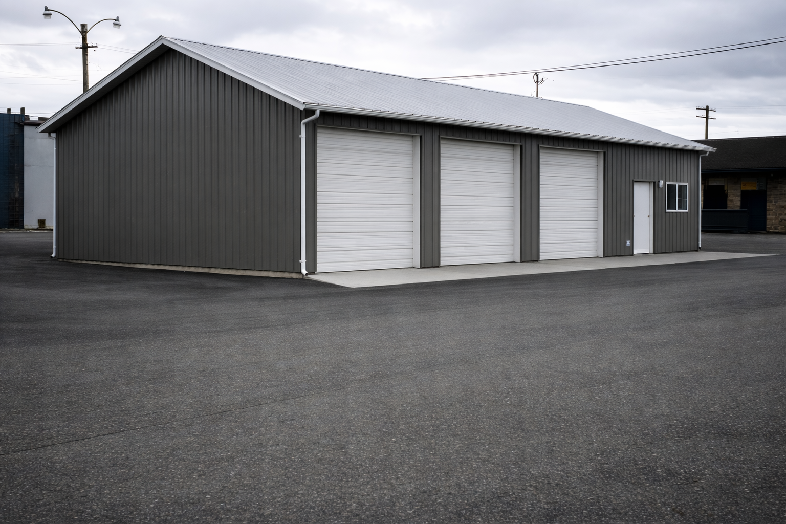 A gray metal storage building with three white garage doors and one small white door, situated on an asphalt lot under an overcast sky.