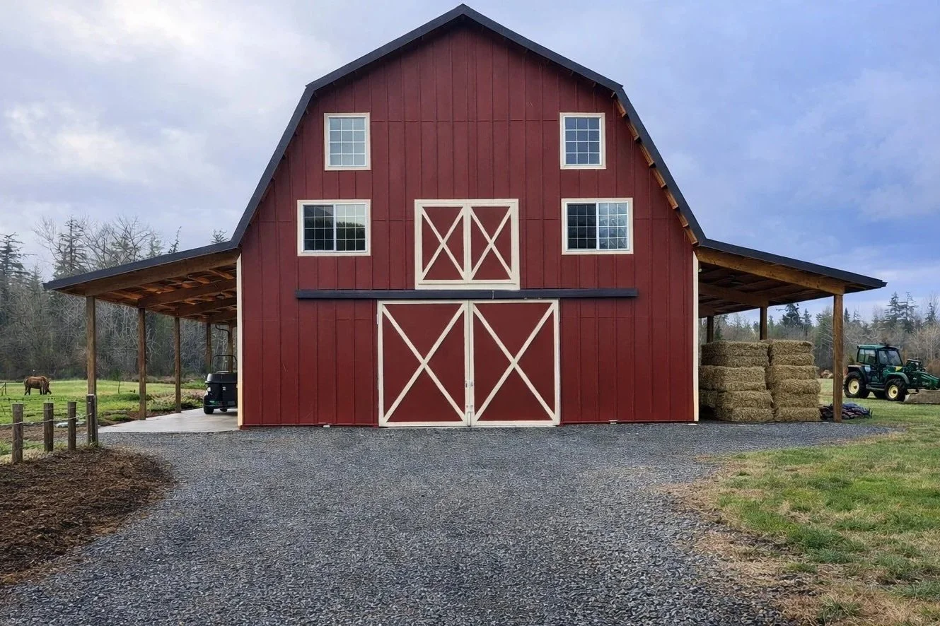 Red barn with white doors and window frames, hay bales on the right, tractor on the right, gravel driveway, farm field and trees in the background.