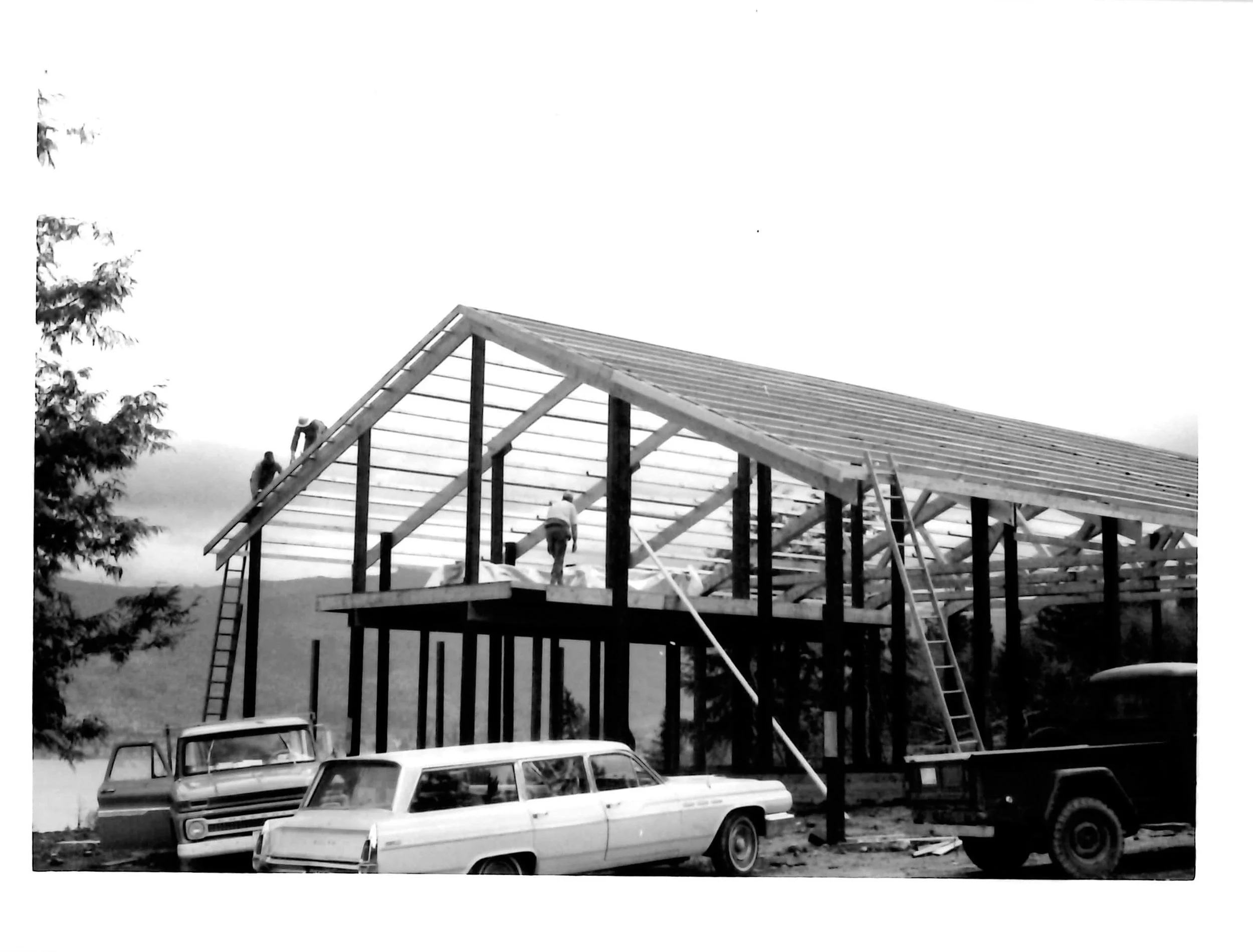 Black and white photo of a pole building under construction, with a wooden framework and workers on site. Several cars are parked in the foreground, including a station wagon and a pickup truck, with a body of water and trees in the background.
