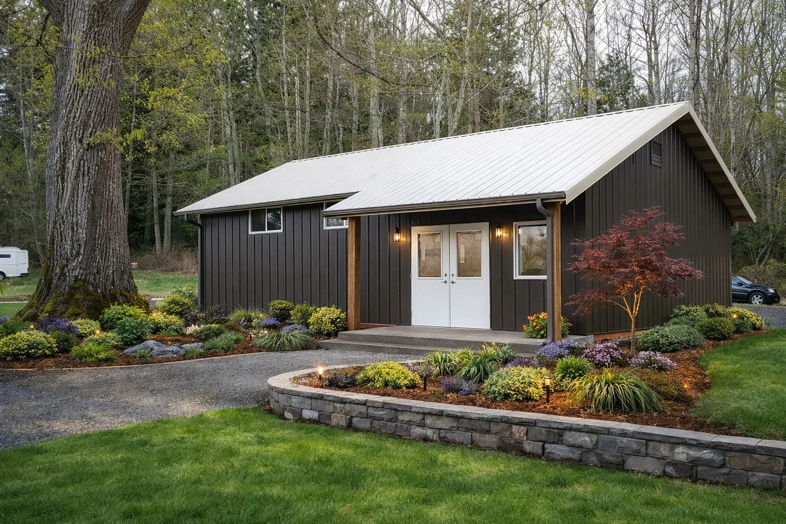 A modern black house with a white metal roof, surrounded by a landscaped garden with colorful flowers, small shrubs, and a young red tree in front. There are trees in the background and a gravel pathway leading to the white double door entrance, which is illuminated by wall-mounted lights.