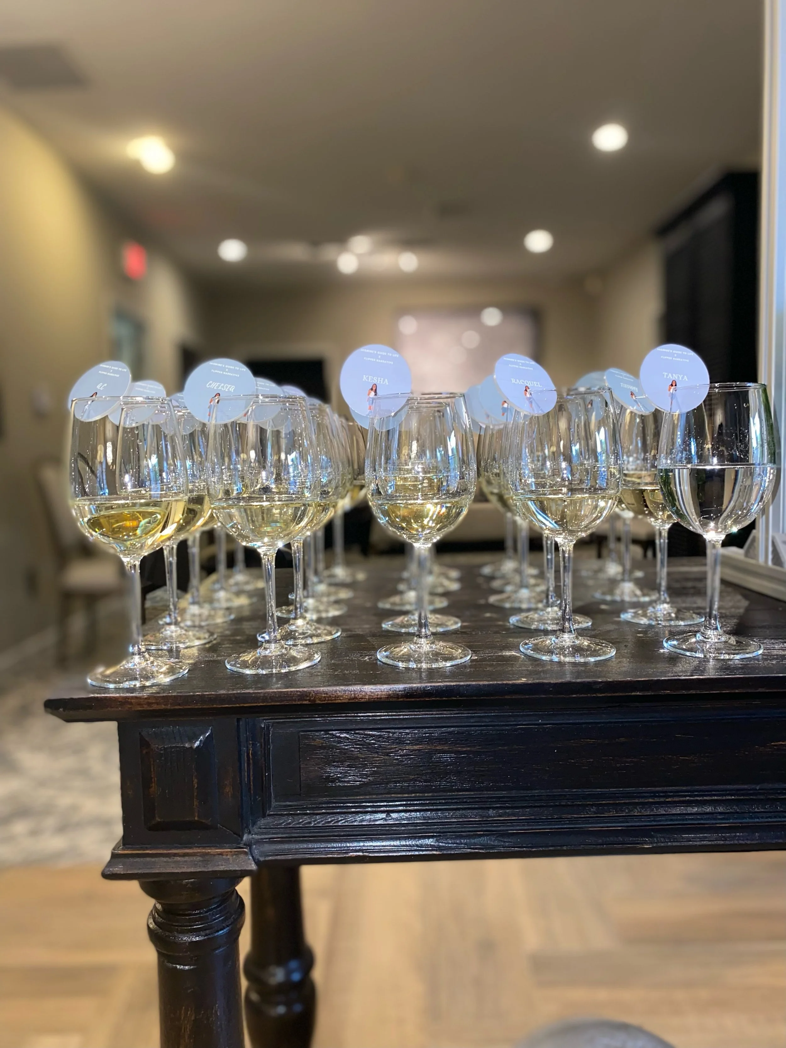 Multiple glasses of white wine on a dark wooden table, each with a small round label with names like Keisha and Tanya, in an indoor setting with blurred background.