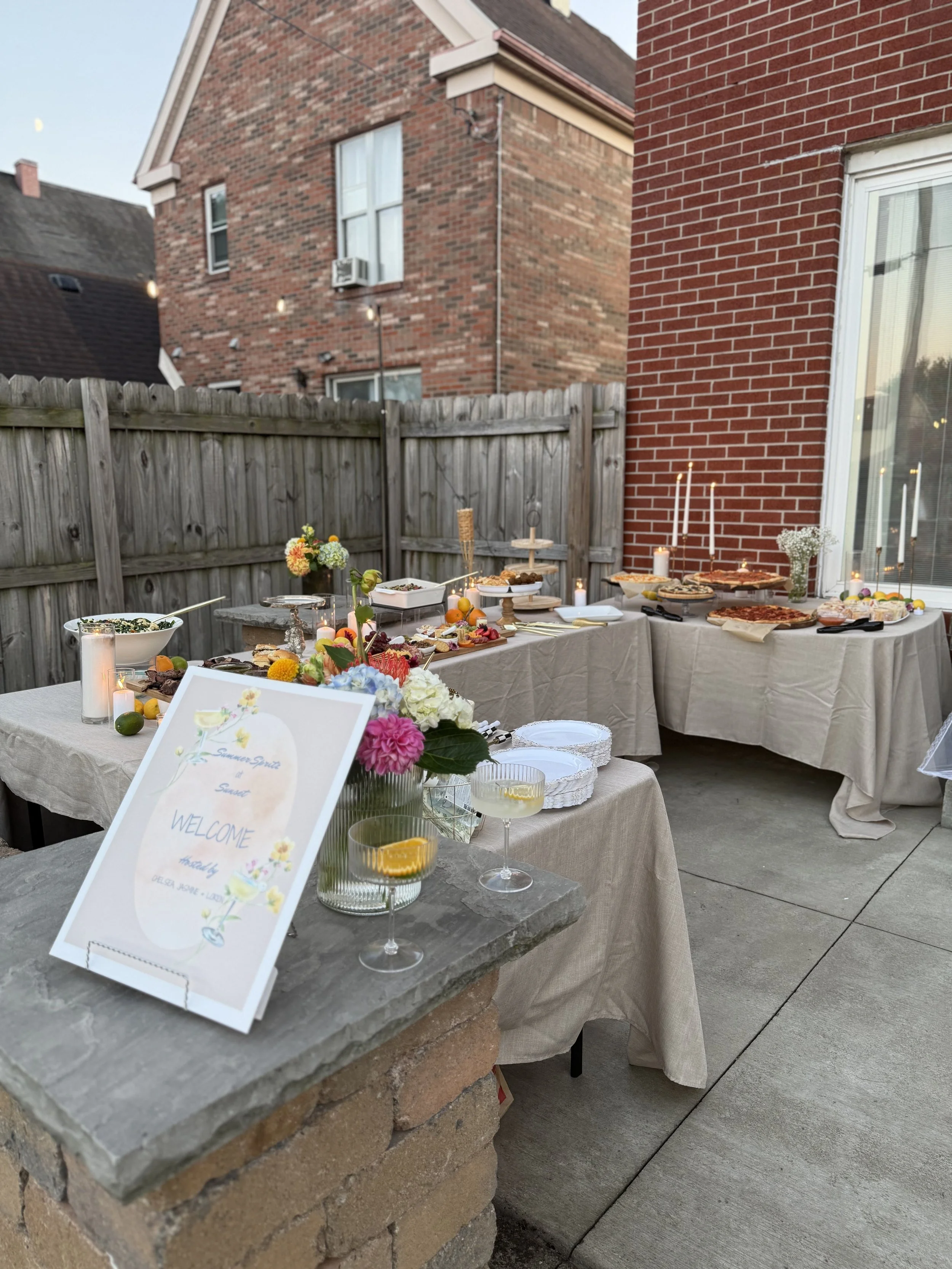 Outdoor buffet table set with various dishes, candles, flowers, and a welcome sign, with brick house and wooden fence in background.