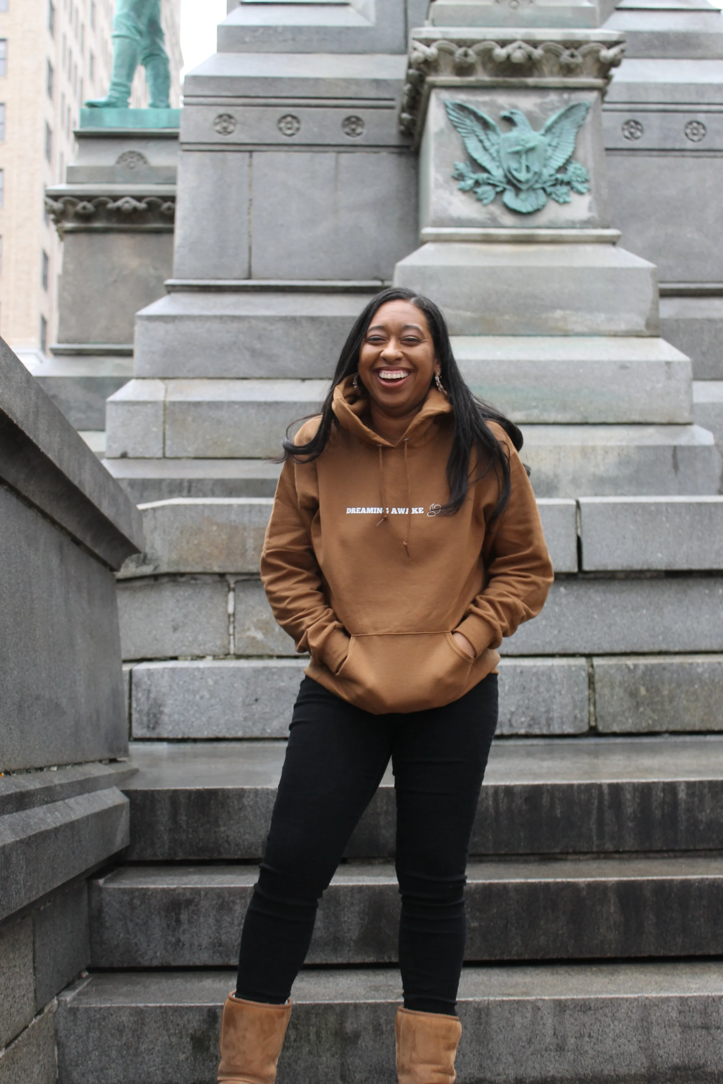 Woman standing on steps in front of a historical monument with a beard and shield emblem, smiling and wearing a brown hoodie, black pants, and tan boots.