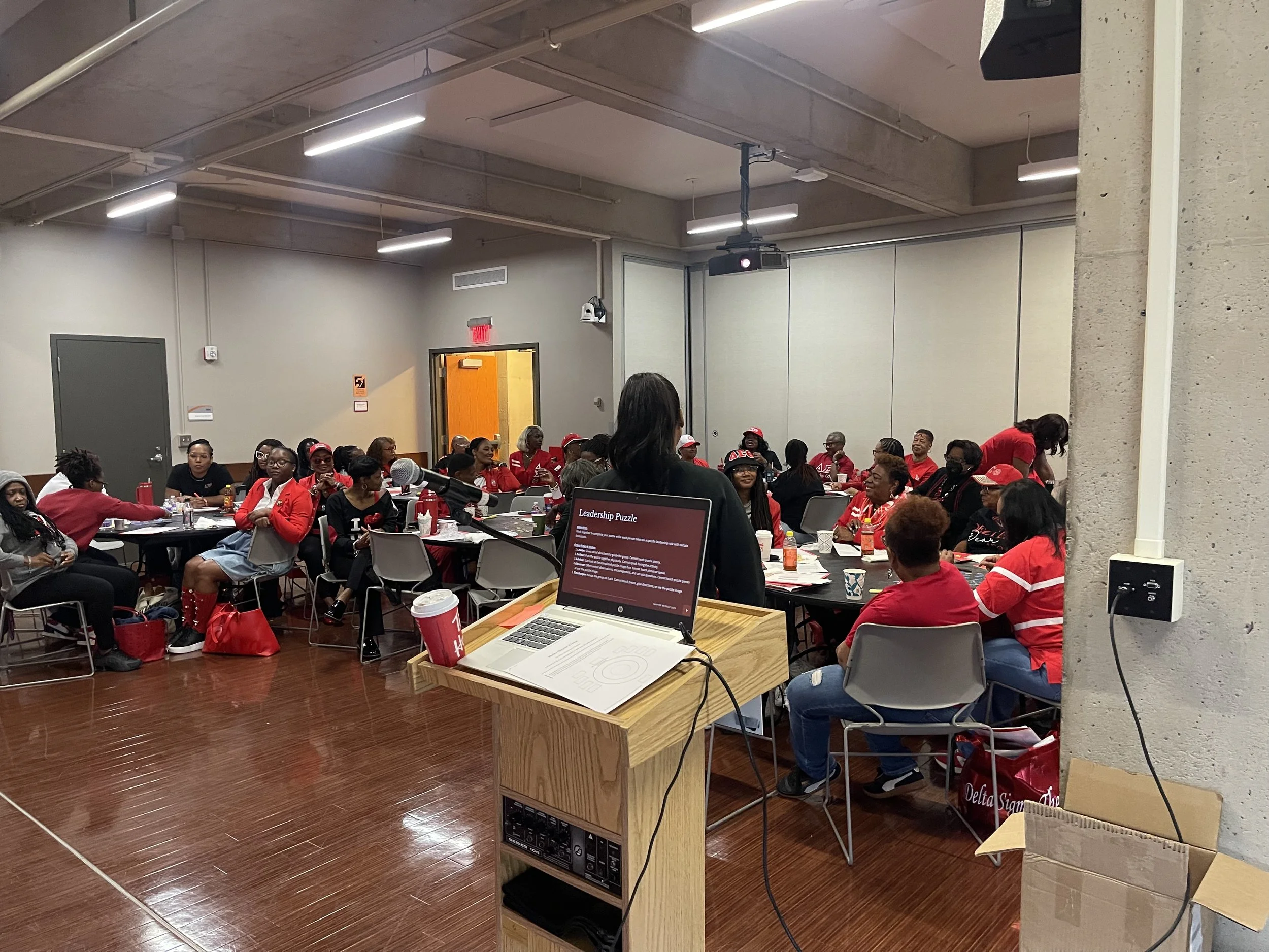 A diverse group of women attending a presentation or workshop in a conference room, seated at round tables, facing a speaker at a podium with a laptop.