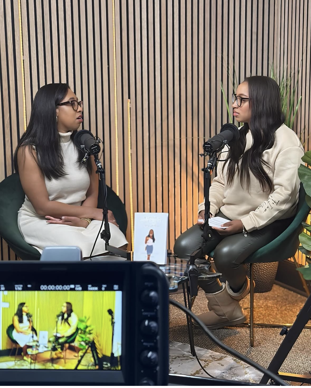 Two women with glasses sitting across from each other, recording a podcast in a studio with a wood-paneled wall background, microphones, and recording equipment.