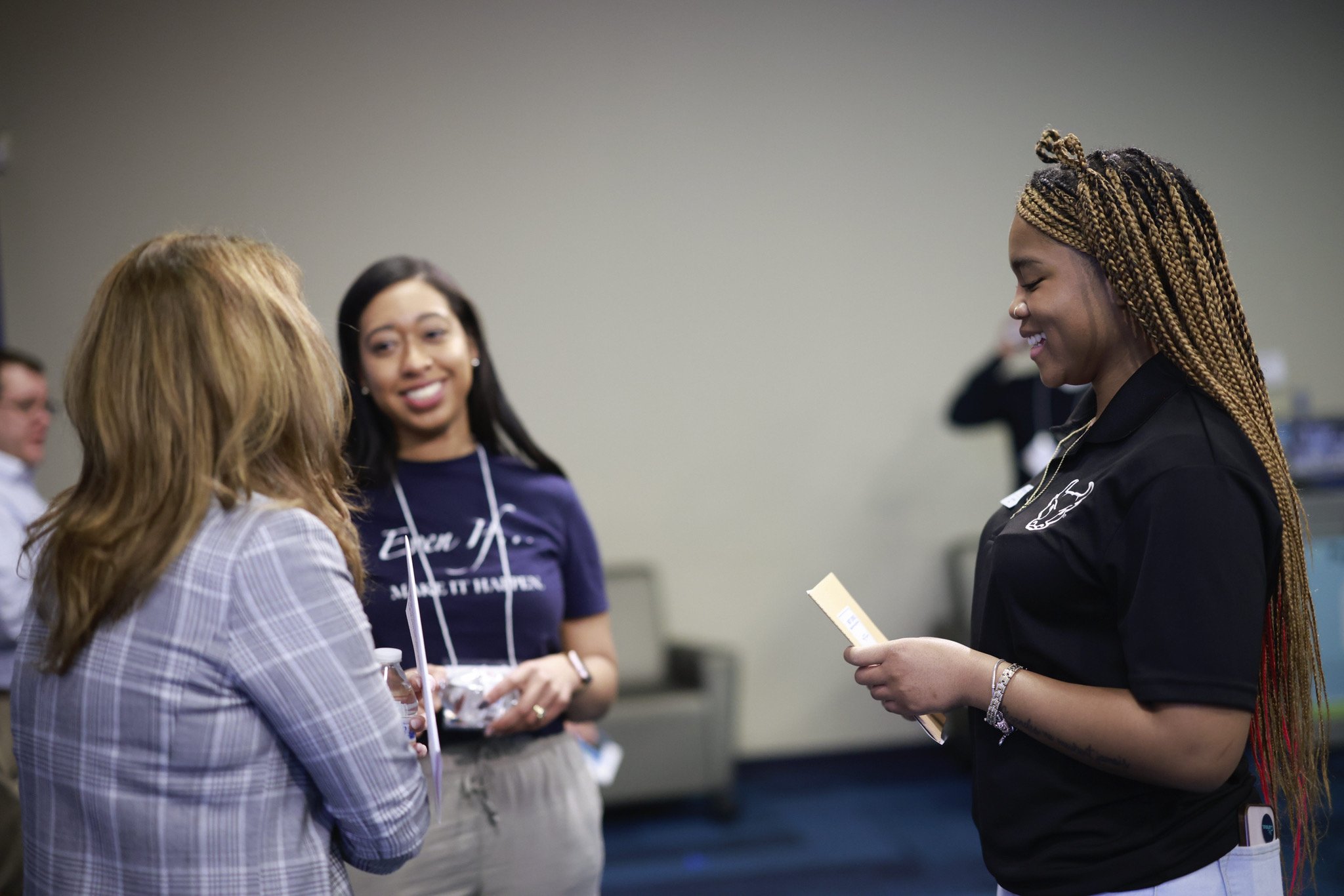 Three women standing and talking at an indoor event. One woman is smiling while looking at a brochure or paper. The women are dressed in casual to business casual attire.