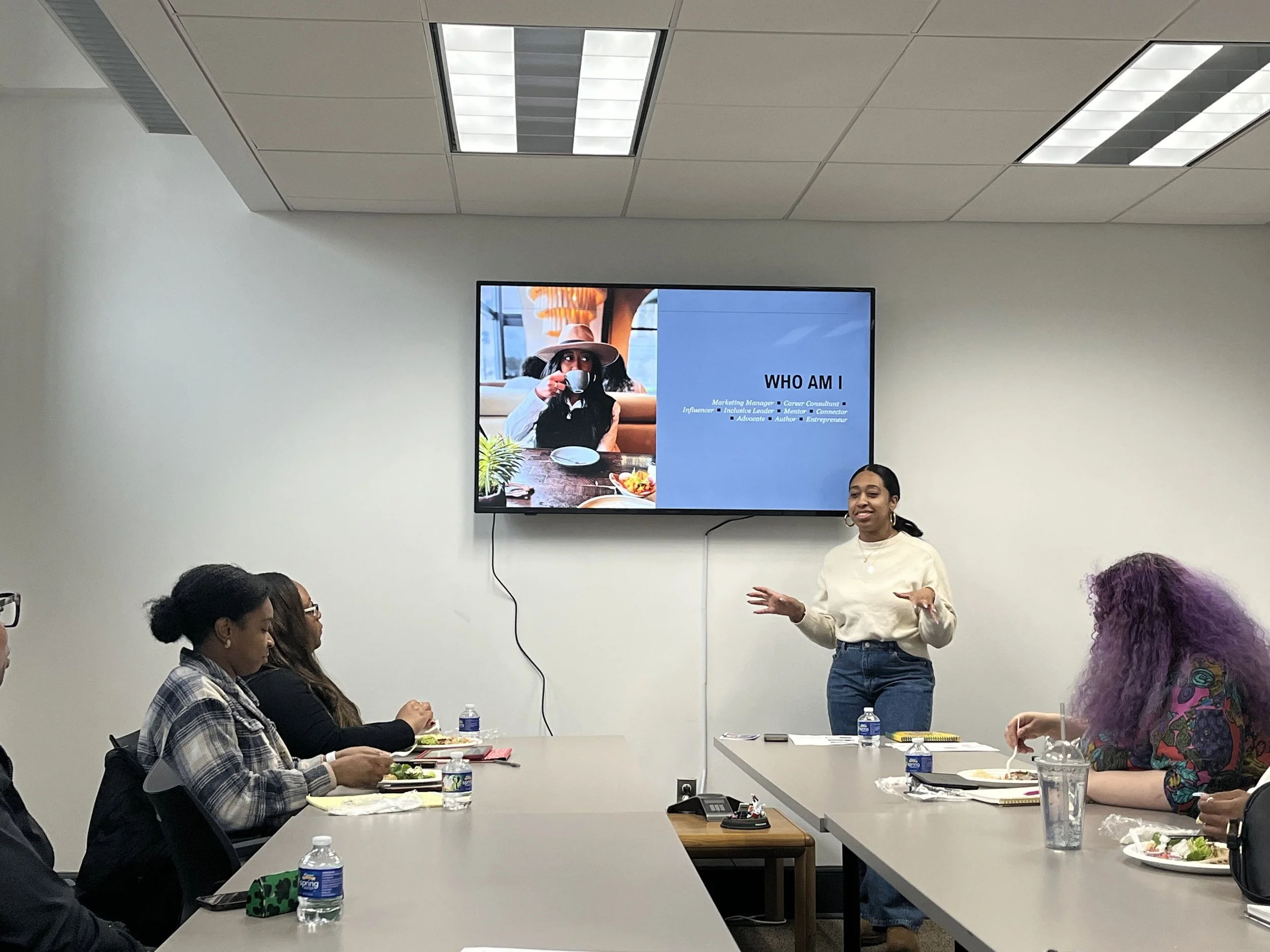 A woman is giving a presentation in a conference room with four people seated at a table. The slide on the screen shows a person drinking coffee and the text 'WHO AM I'.