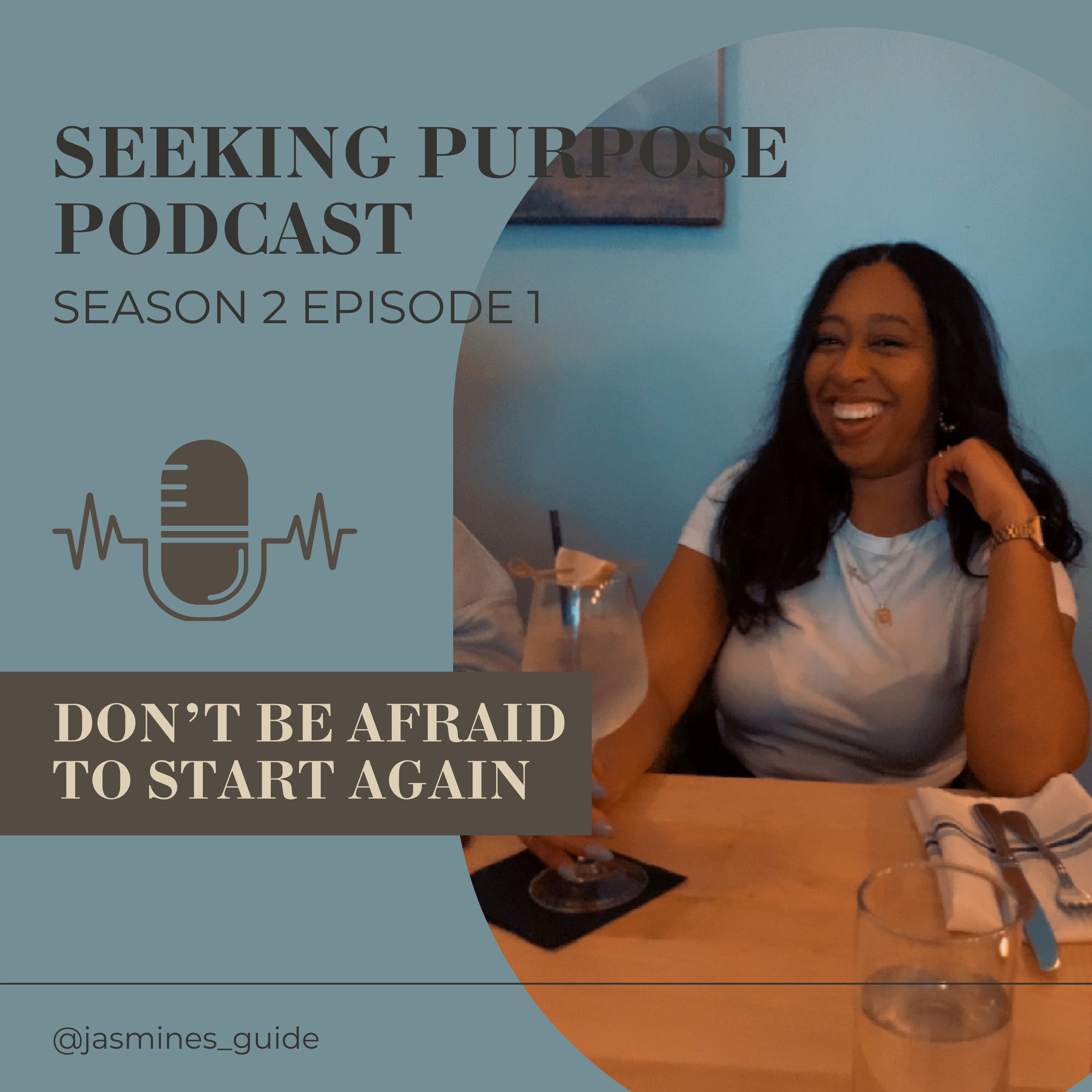 A woman with long black hair, wearing a white shirt and jewelry, smiling and sitting at a restaurant table with a glass of water, a napkin, and utensils, with a blue wall in the background.