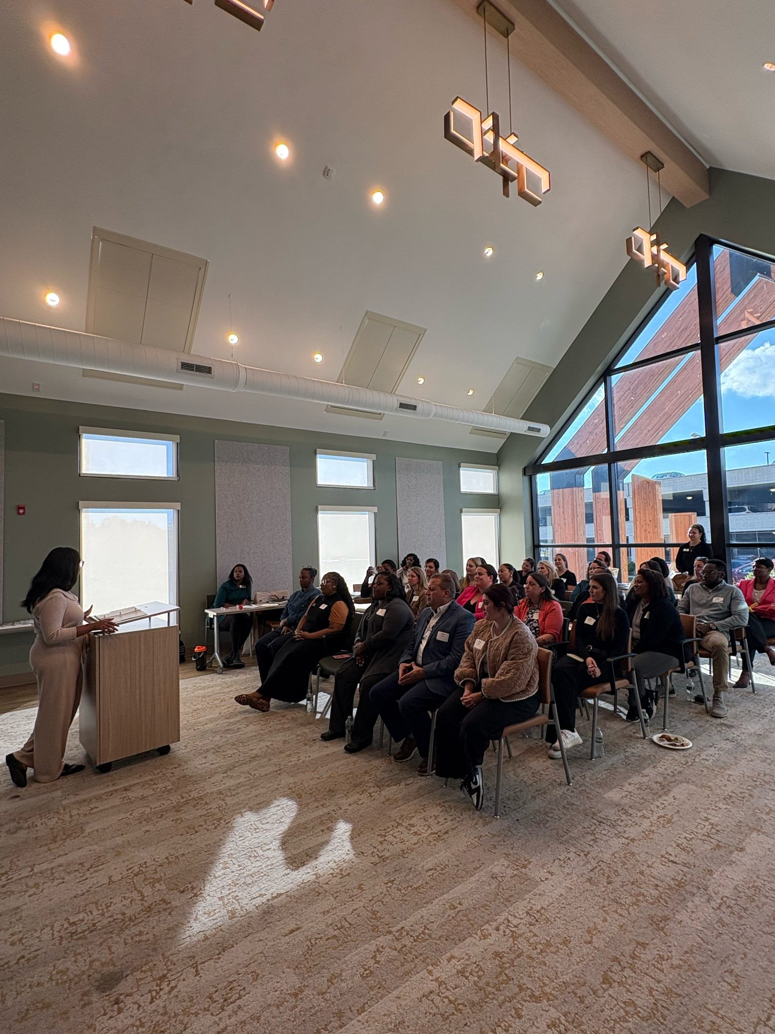 A woman is giving a presentation to a seated audience in a modern conference room with large windows.