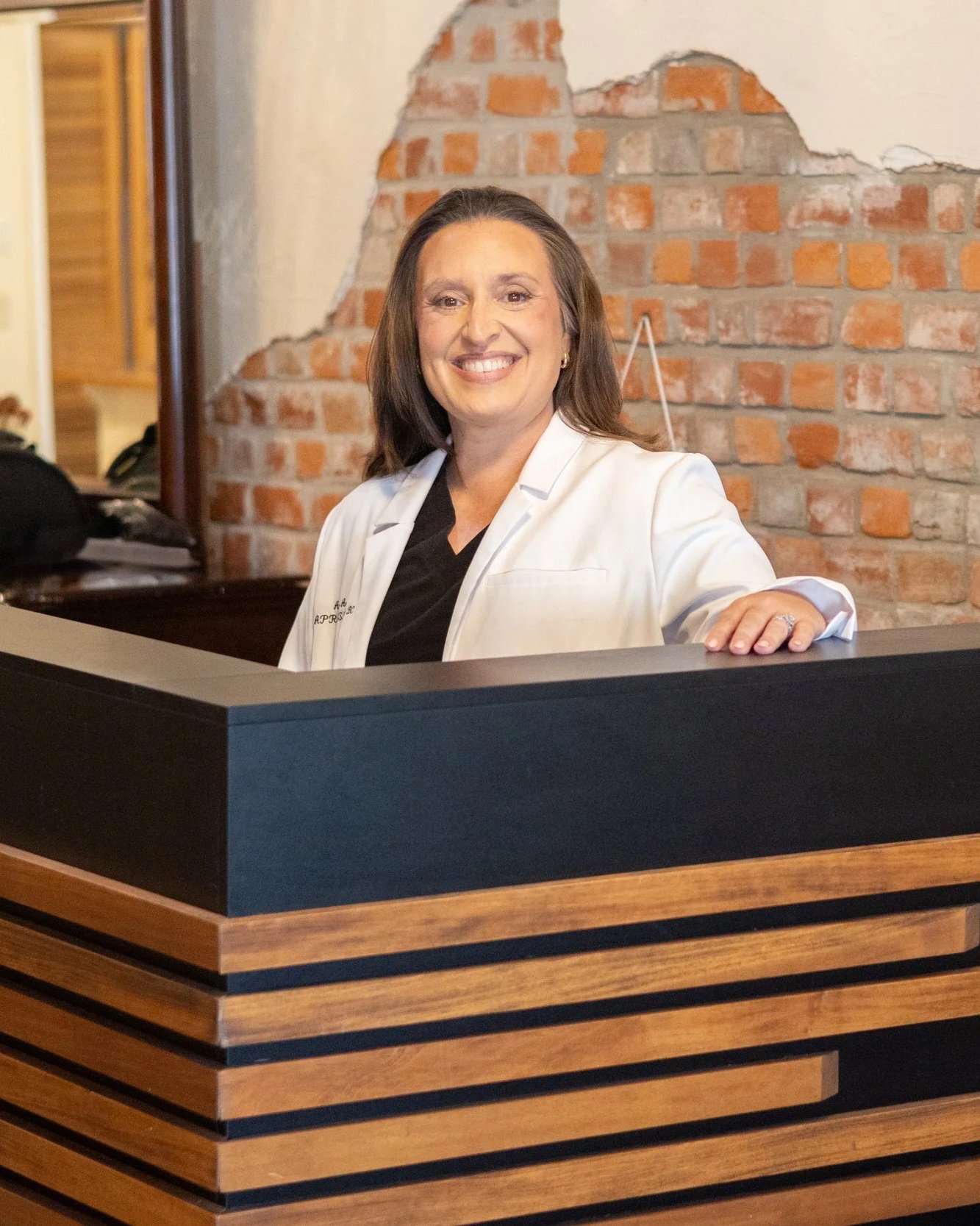 Angela, co-founder of Verity Health & Aesthetics, smiling in black medical scrubs and lab coat