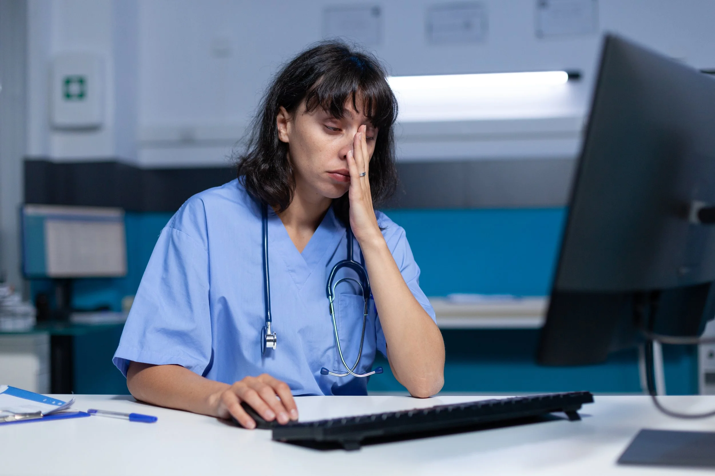 A female healthcare worker in a blue medical uniform is sitting at a desk with her eyes closed and her hand resting on her face, appearing stressed or exhausted, with medical equipment and documents on the desk in a clinical setting.