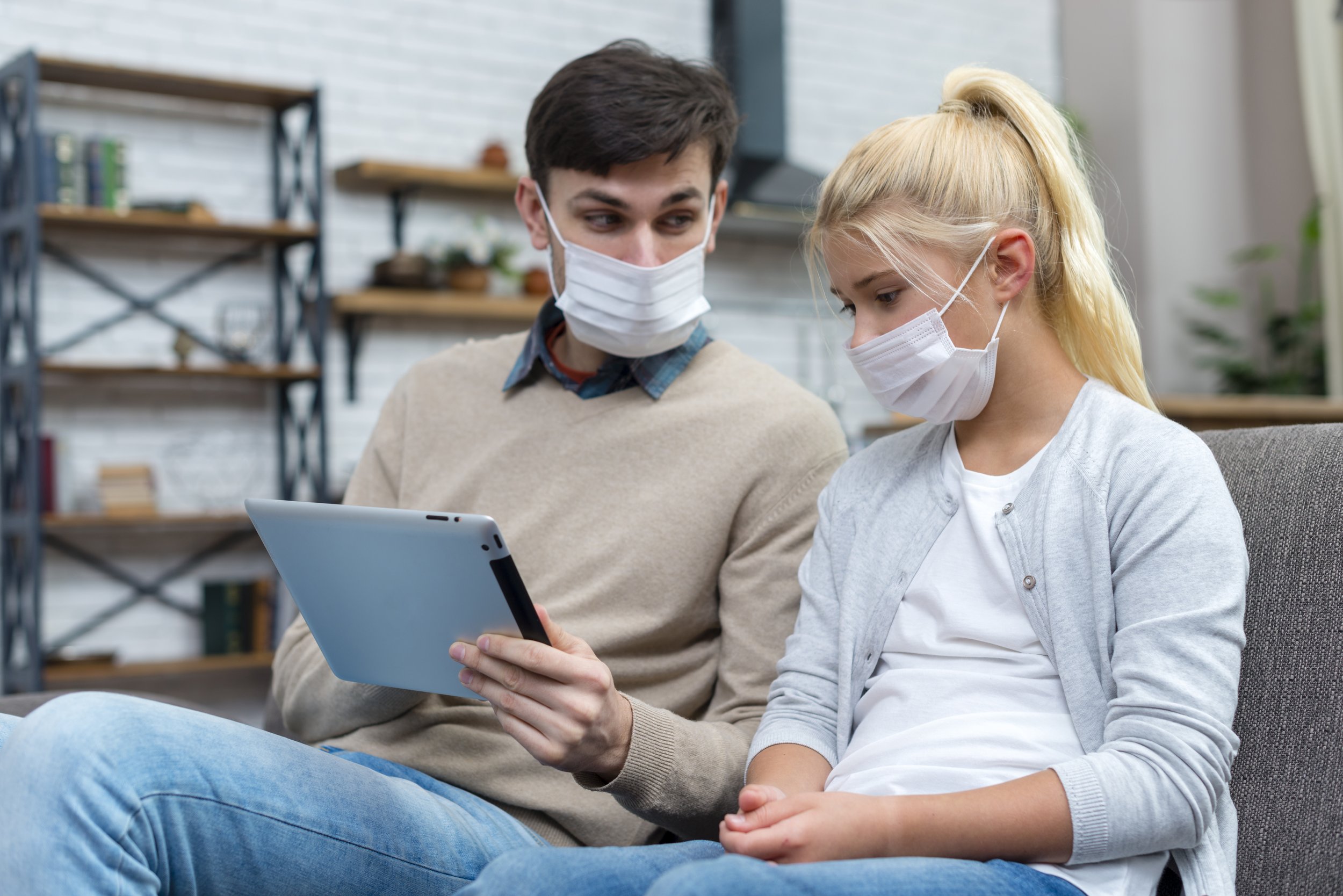 A young man and a young girl sitting on a gray sofa, both wearing white face masks and looking at a tablet device the girl is holding.