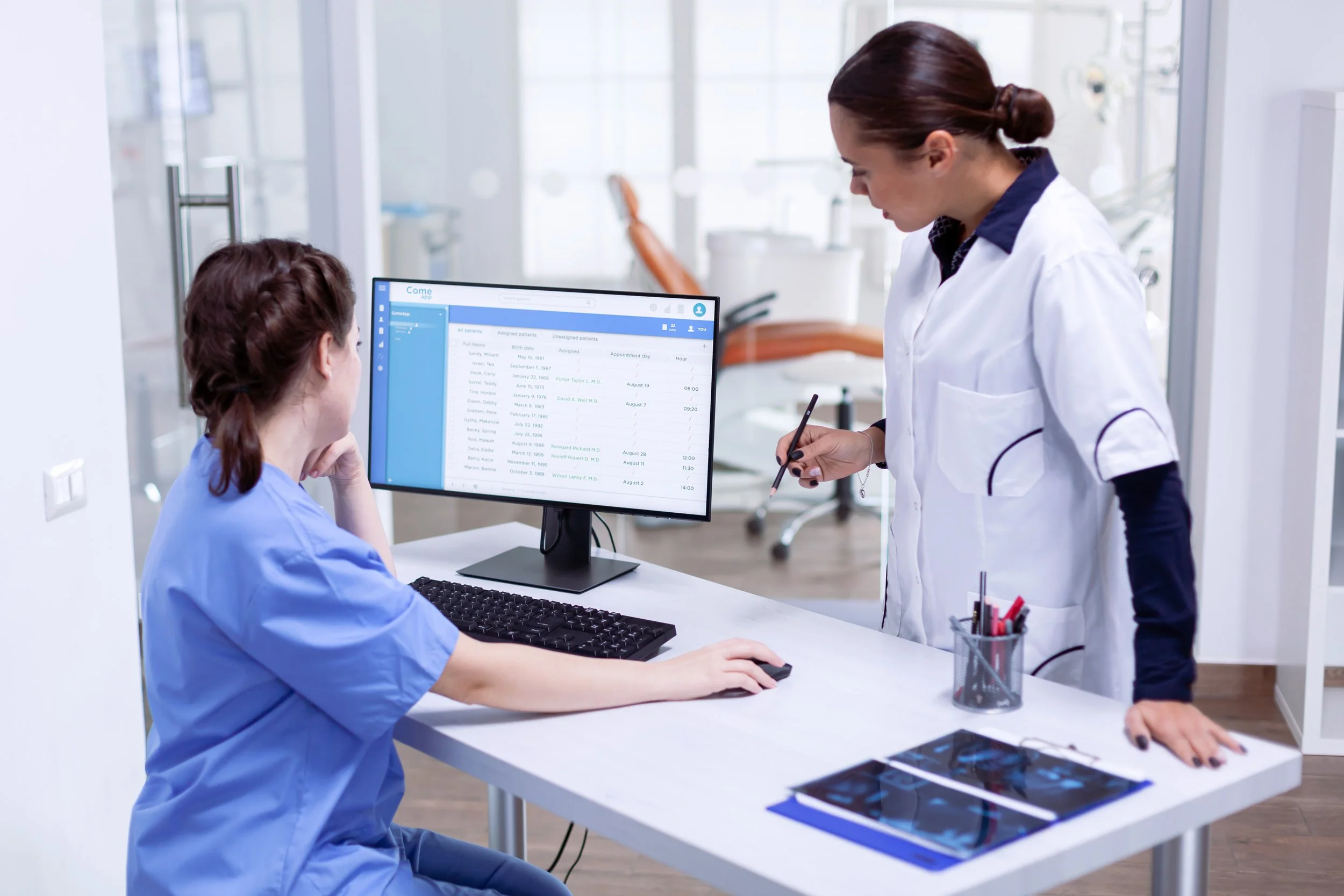 A healthcare professional, likely a nurse or doctor in blue scrubs, sitting at a desk in a medical office or hospital, looking at a computer monitor displaying patient information. A female colleague in a white lab coat is standing beside her, writing on a clipboard or notepad.