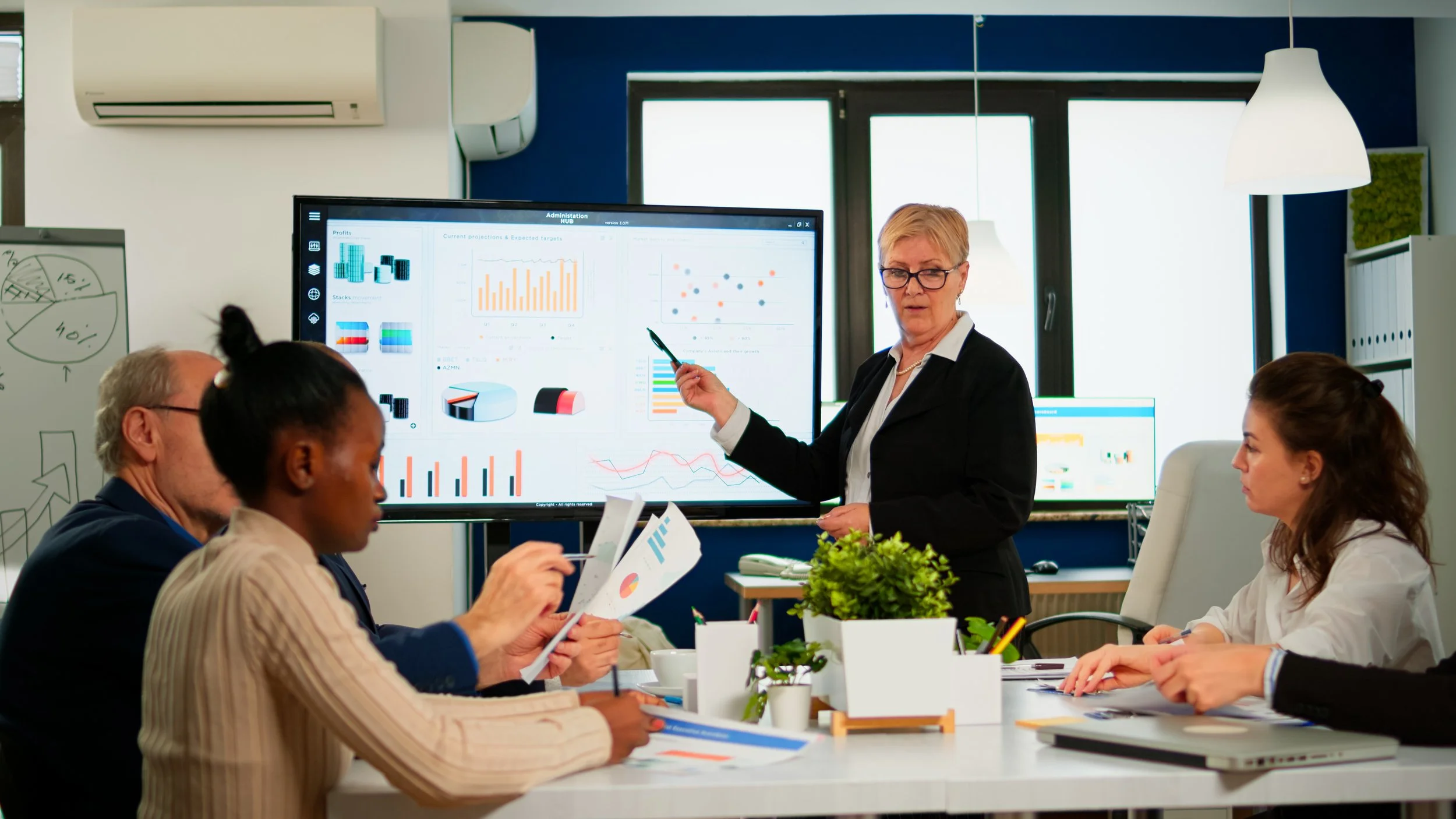 A business presentation meeting with a woman pointing at a large monitor displaying charts and graphs. Four people are seated at the table, reviewing documents, with a whiteboard and windows in the background.