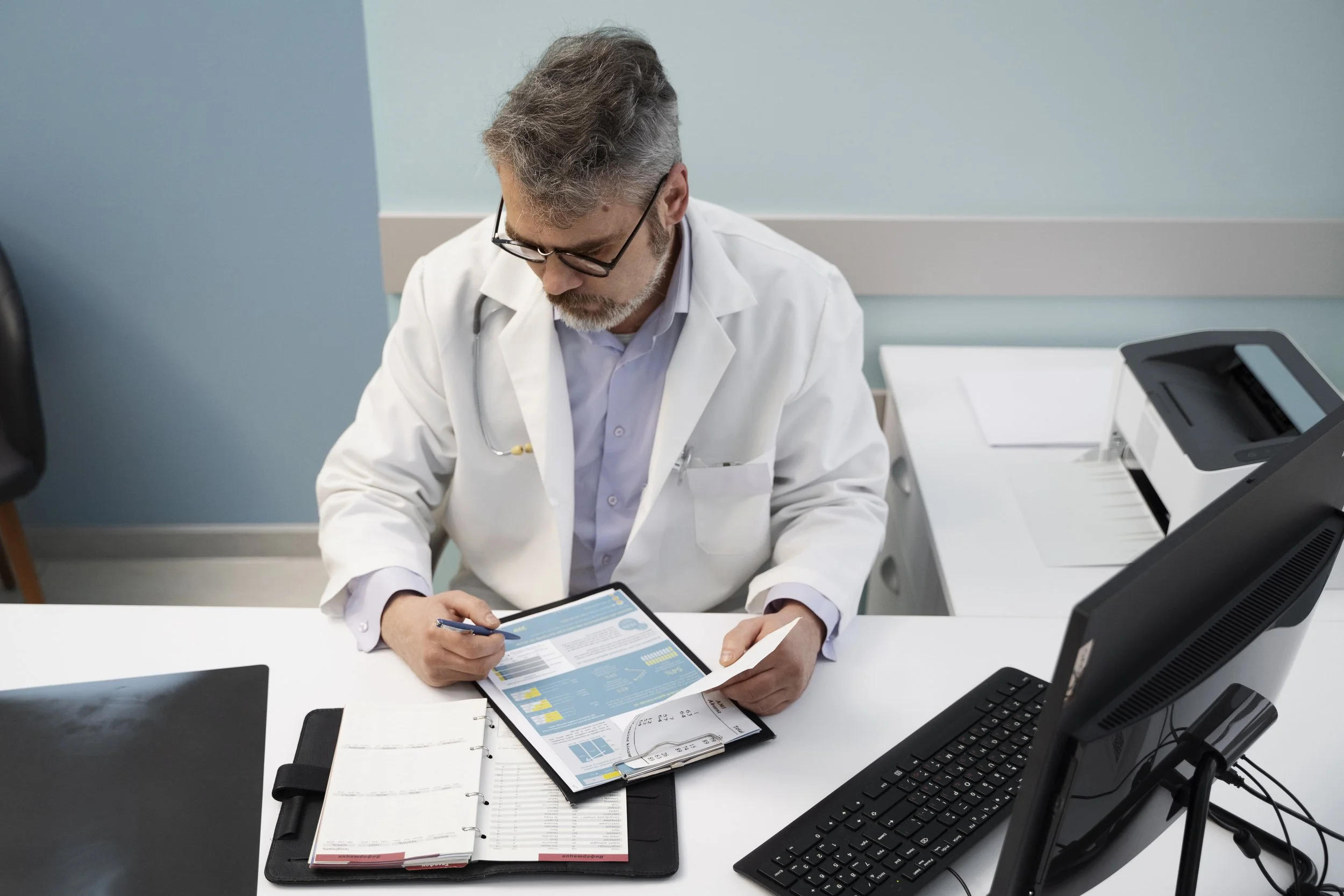 A male doctor with gray hair and glasses wearing a white lab coat, sitting at a desk in a medical office, looking at a clipboard with charts and documents, with a computer and printer on the desk.