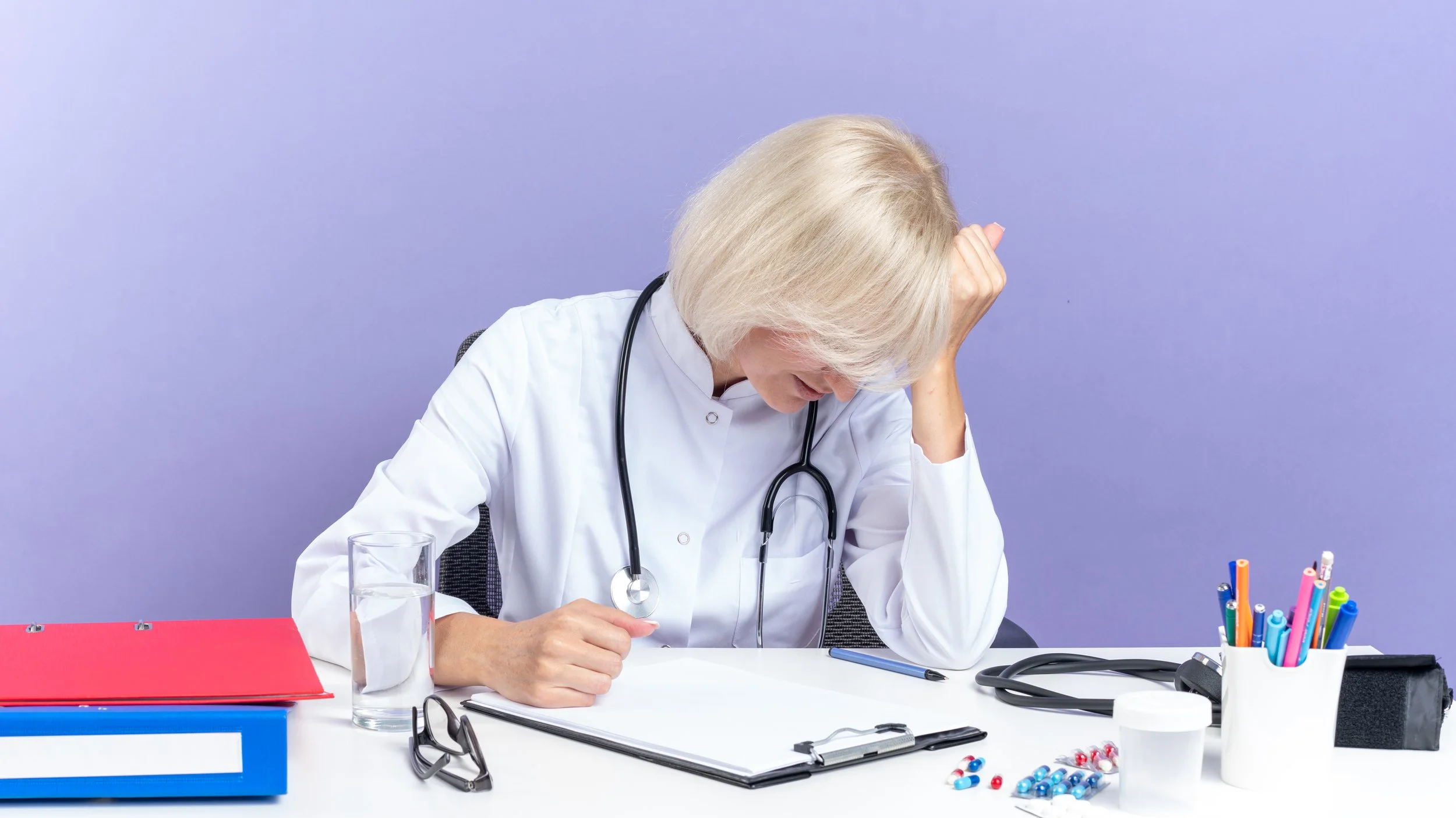 A doctor with gray hair, wearing a white coat and stethoscope, sitting at a desk with a distressed expression, hand on forehead, surrounded by medical supplies and colorful pens against a purple background.