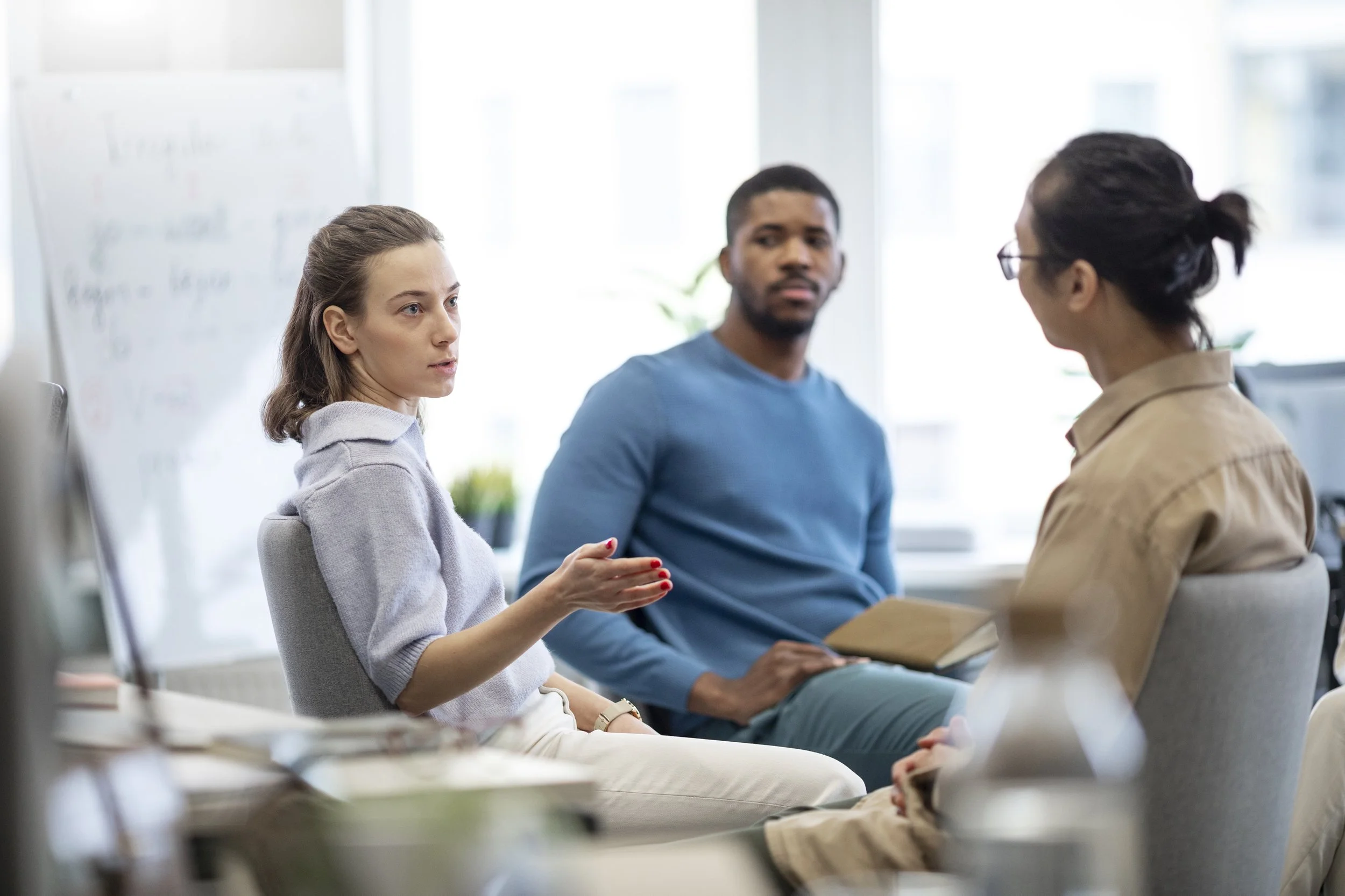 Three people engaged in a discussion in an office, with a woman on the left speaking and a man and woman listening.